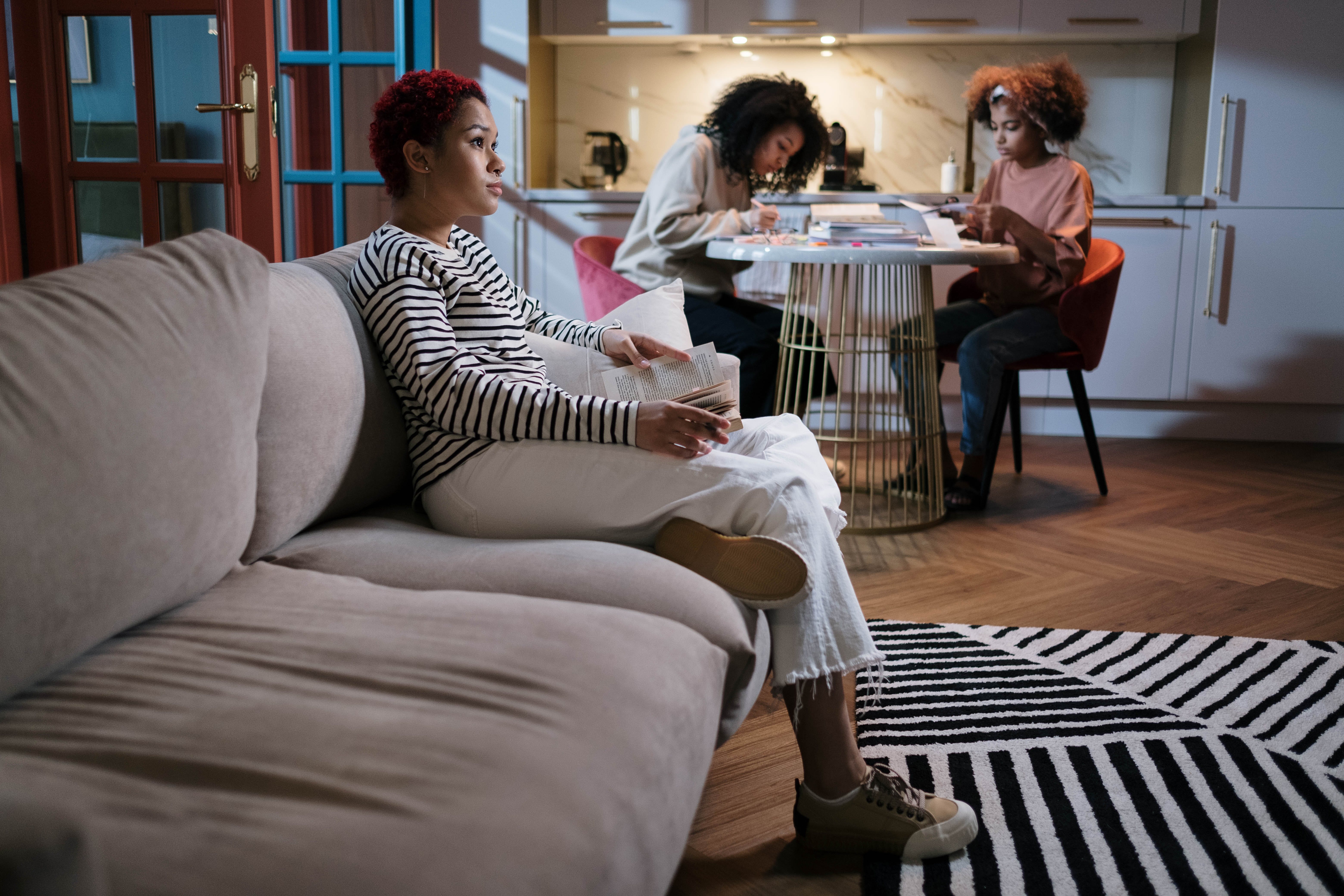 A person reads on a sofa while two others work at a table in a modern kitchen-living space.