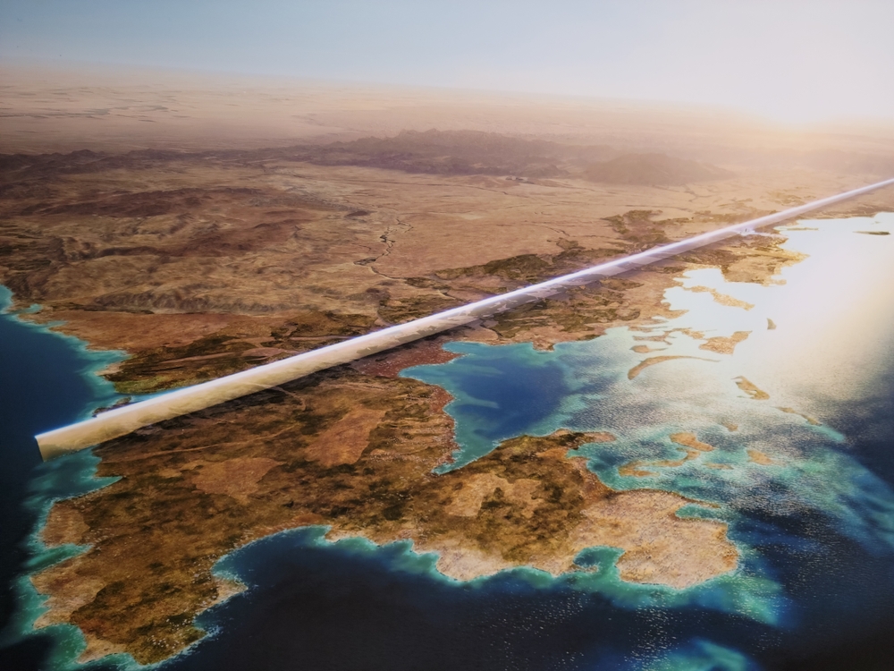 A futuristic linear structure spans across a desert landscape and coastal waters at sunset.
