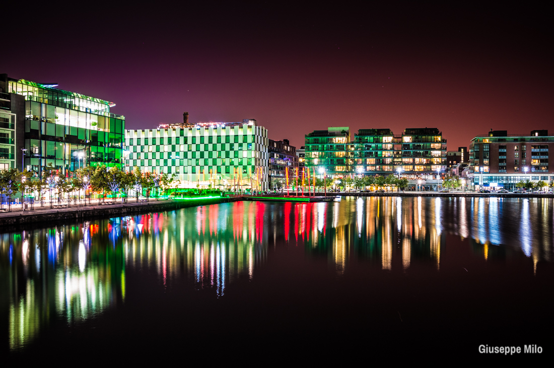 A vibrant waterfront cityscape at night, with colorful reflections and modern architecture.