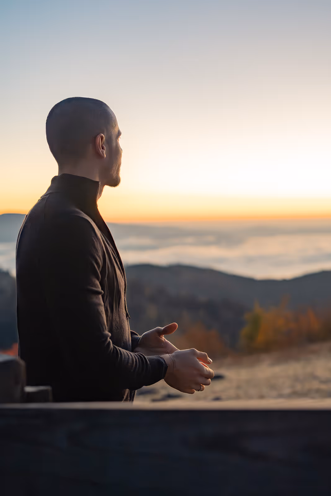 Ein Mann mit kurzem Haar und schwarzem Pullover schaut bei Sonnenaufgang auf Berge und Nebel.