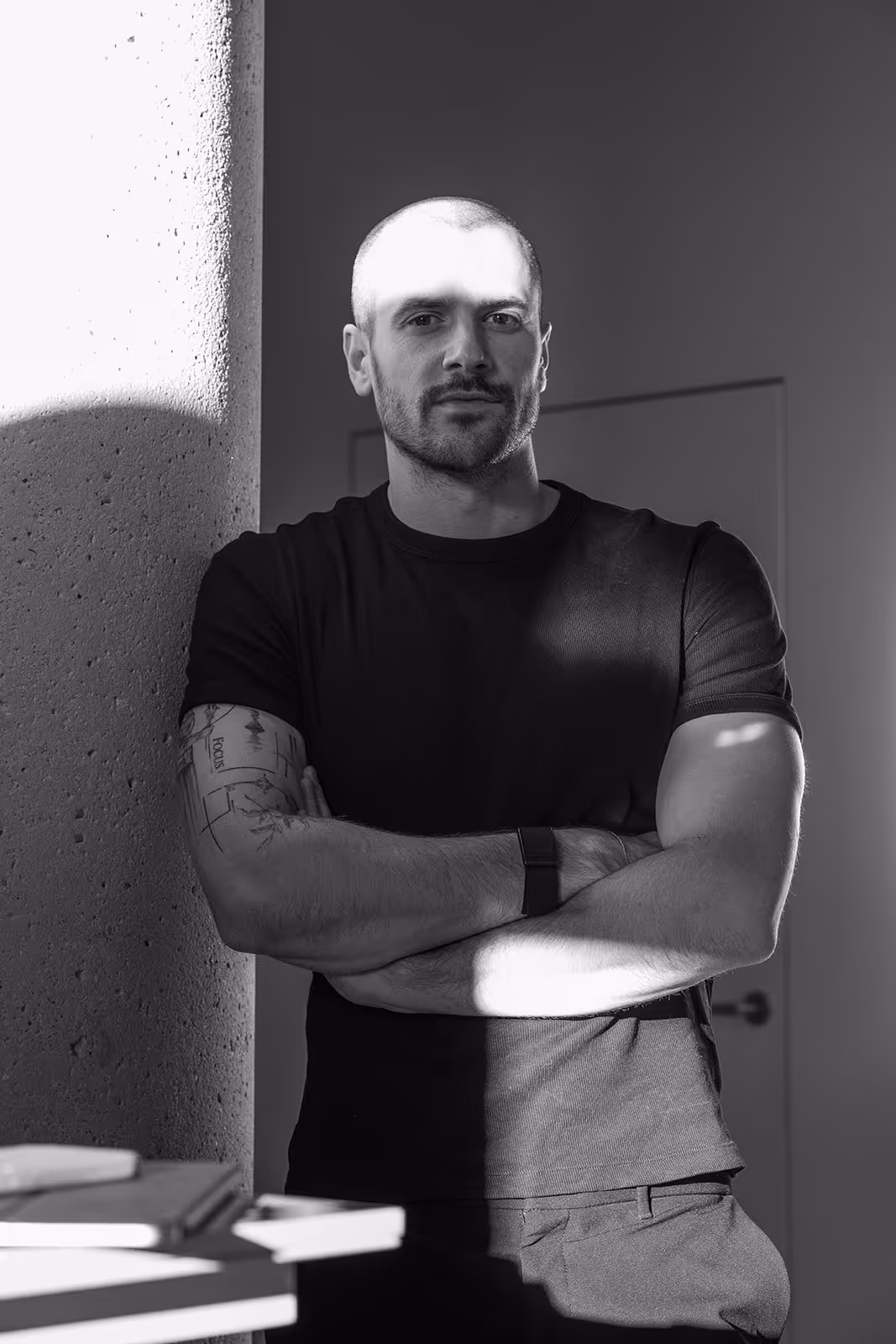 Schwarz-weißes Porträt eines Mannes mit kurzem Haar und Tattoos an einem strukturierten Betonpfeiler, Arme verschränkt, mit Schatten auf Gesicht und Oberkörper.