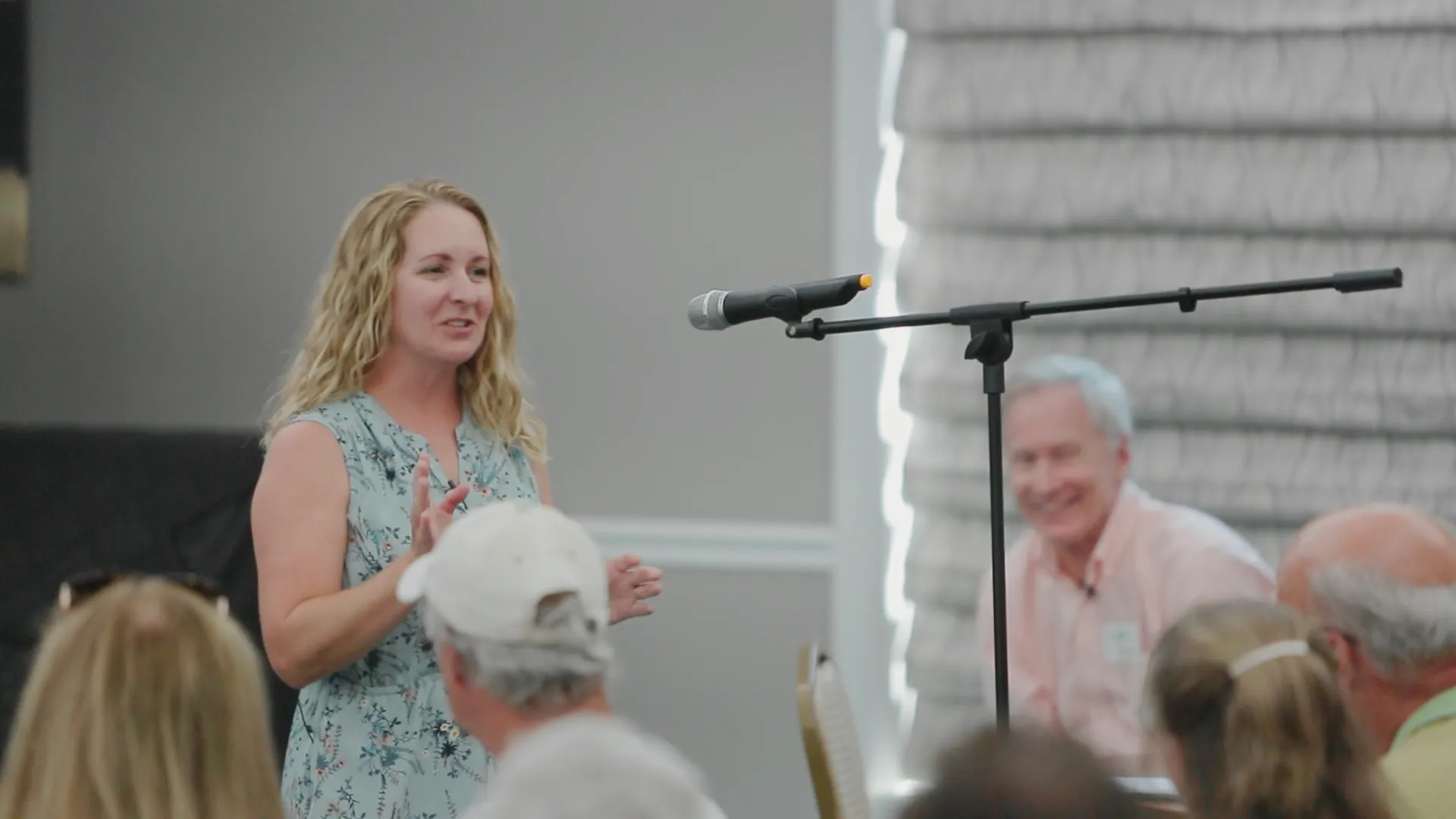 Woman speaking to an audience with a microphone on a stand in front of her, while a man smiles seated in the background.
