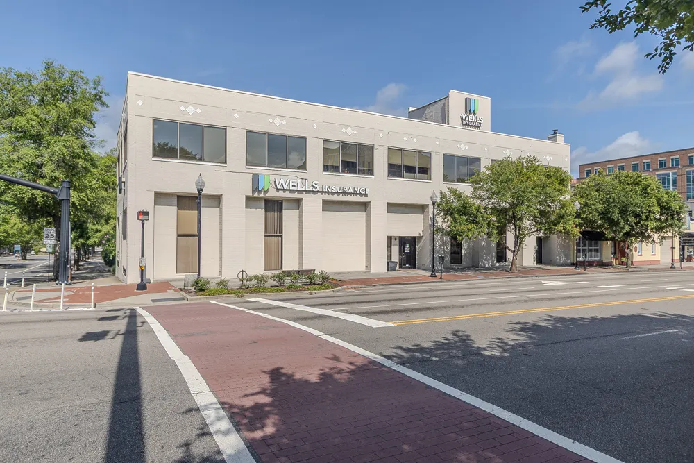Two-story beige building housing Wells Insurance on a sunny day at a street intersection with trees nearby.