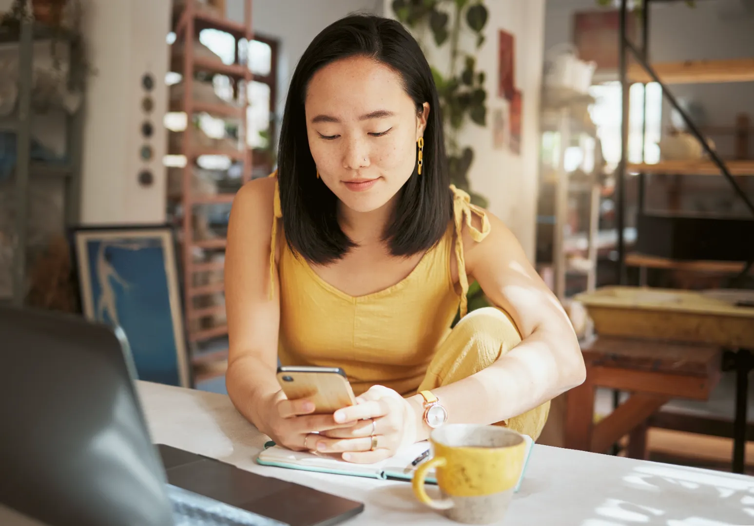 Young woman in yellow outfit sitting at a table, looking at her smartphone with a laptop, notebook, and yellow mug nearby.