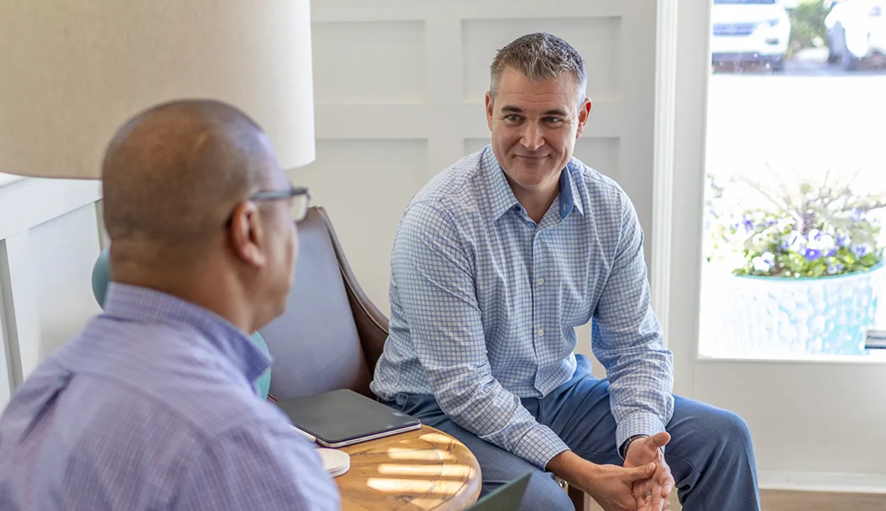 Two men engaged in a friendly conversation in a bright room with a lamp and a wooden table between them.