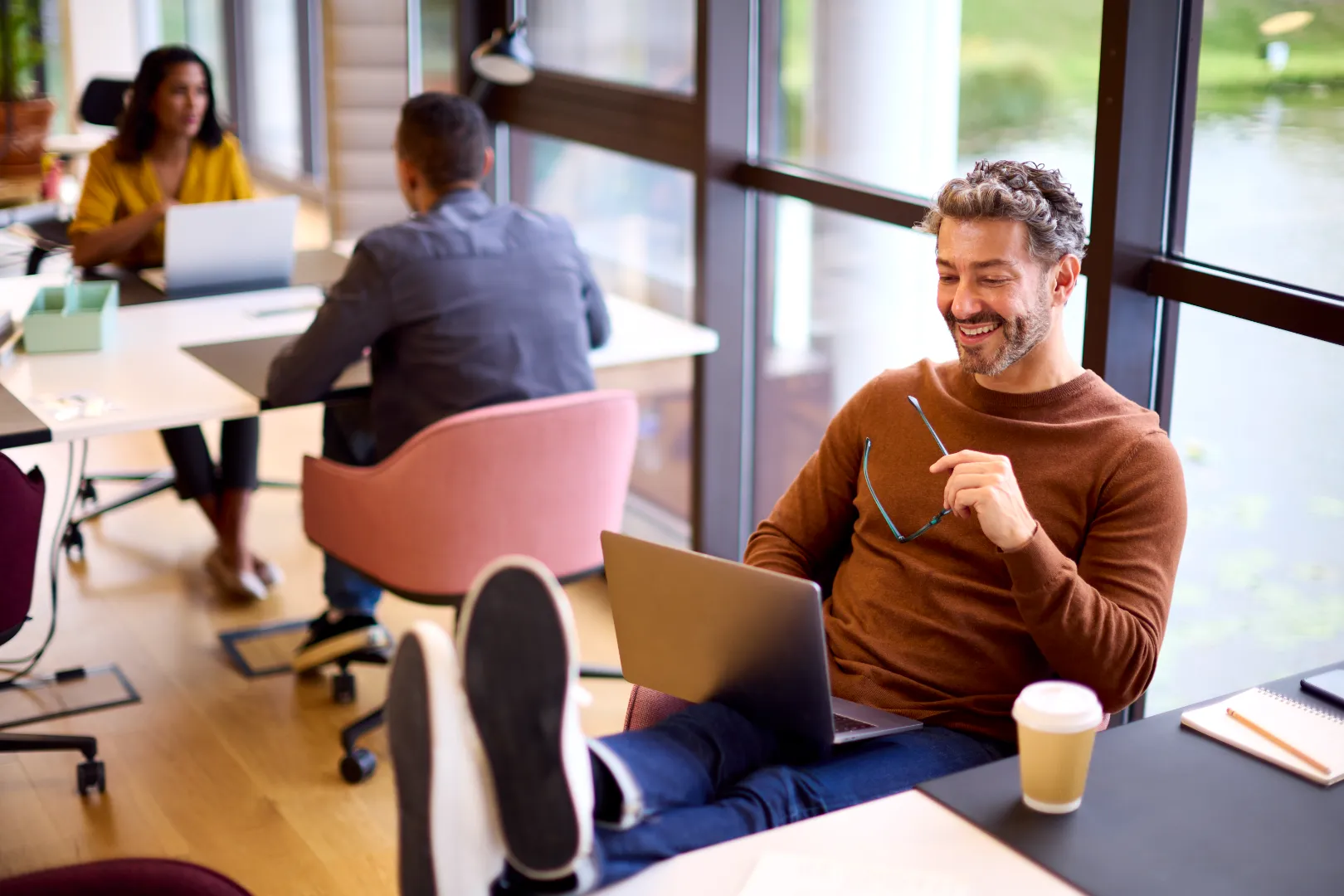 Man in brown sweater sitting with feet up on desk, holding glasses and smiling at laptop in bright office.