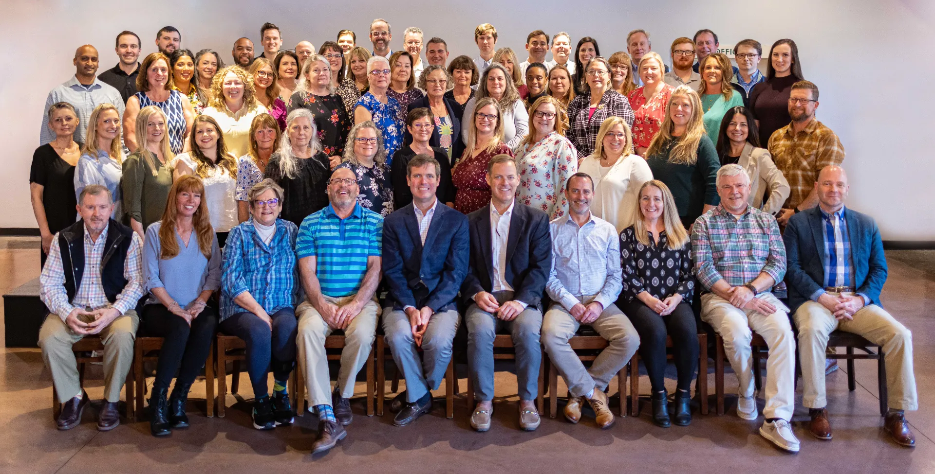 Large group portrait of diverse adults arranged in three rows in a well-lit indoor space.