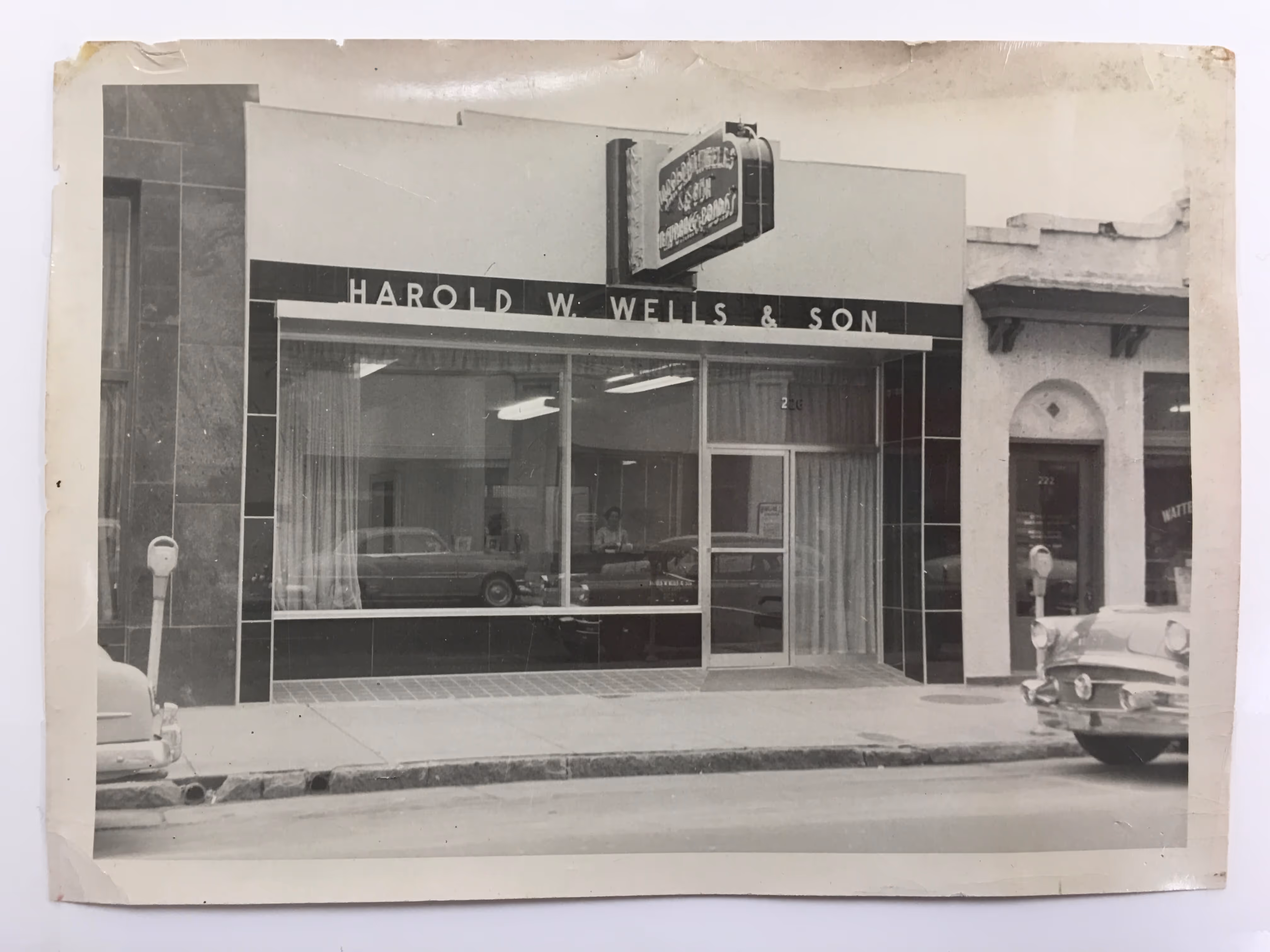 Black-and-white photo of a vintage storefront with the sign 'Harold W. Wells & Son' above large display windows and a door, with classic cars parked on the street.