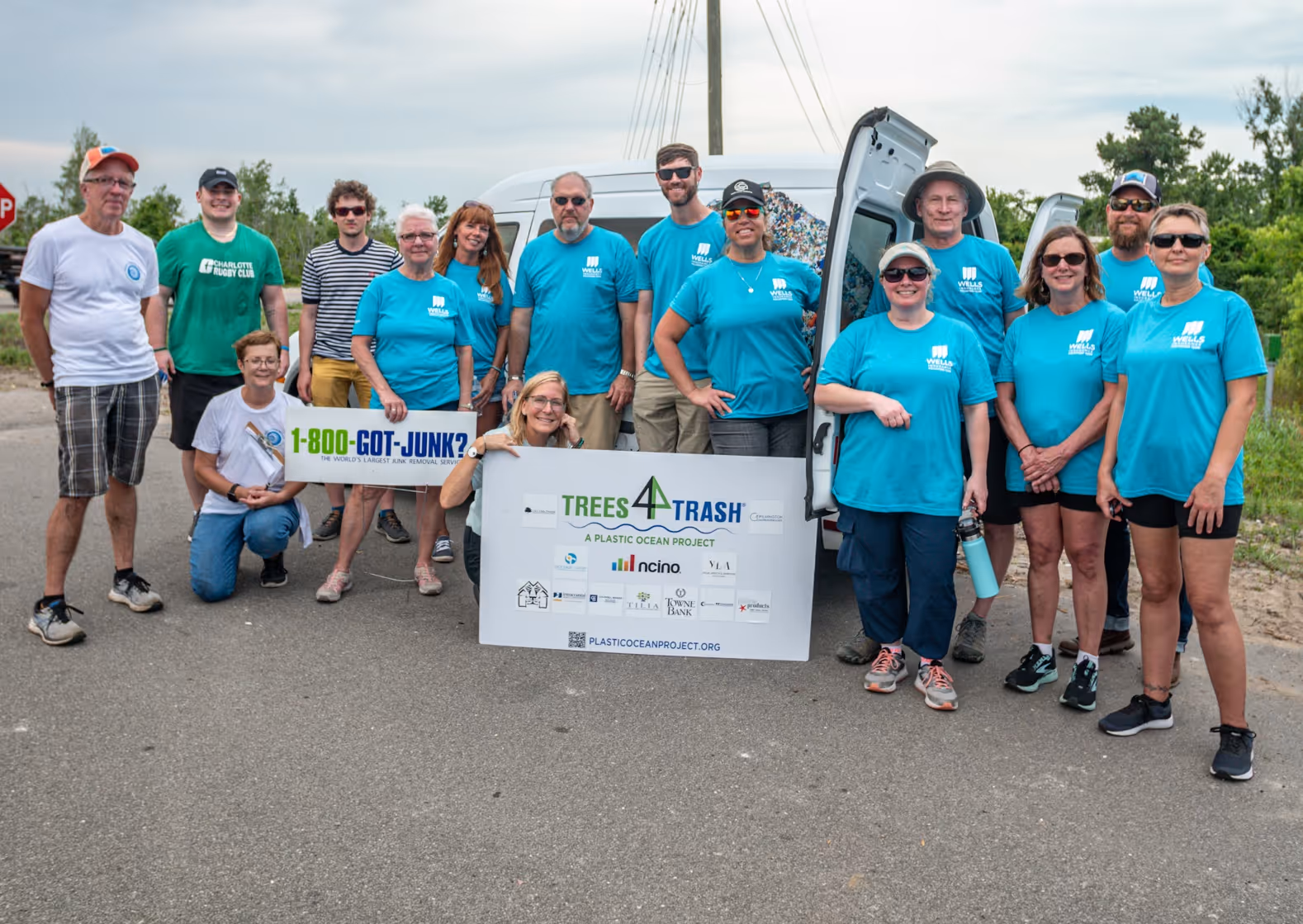 Group of people standing and kneeling outdoors wearing matching blue shirts holding signs for Trees 4 Trash plastic ocean cleanup project.