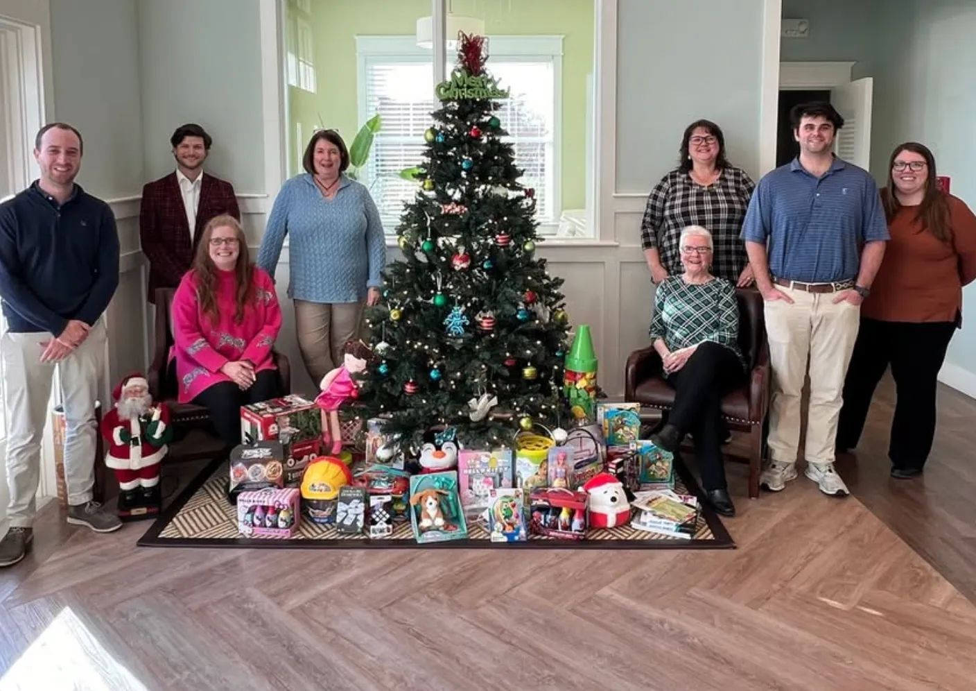 Nine people posing around a decorated Christmas tree with various toys and gifts arranged in front of it indoors.