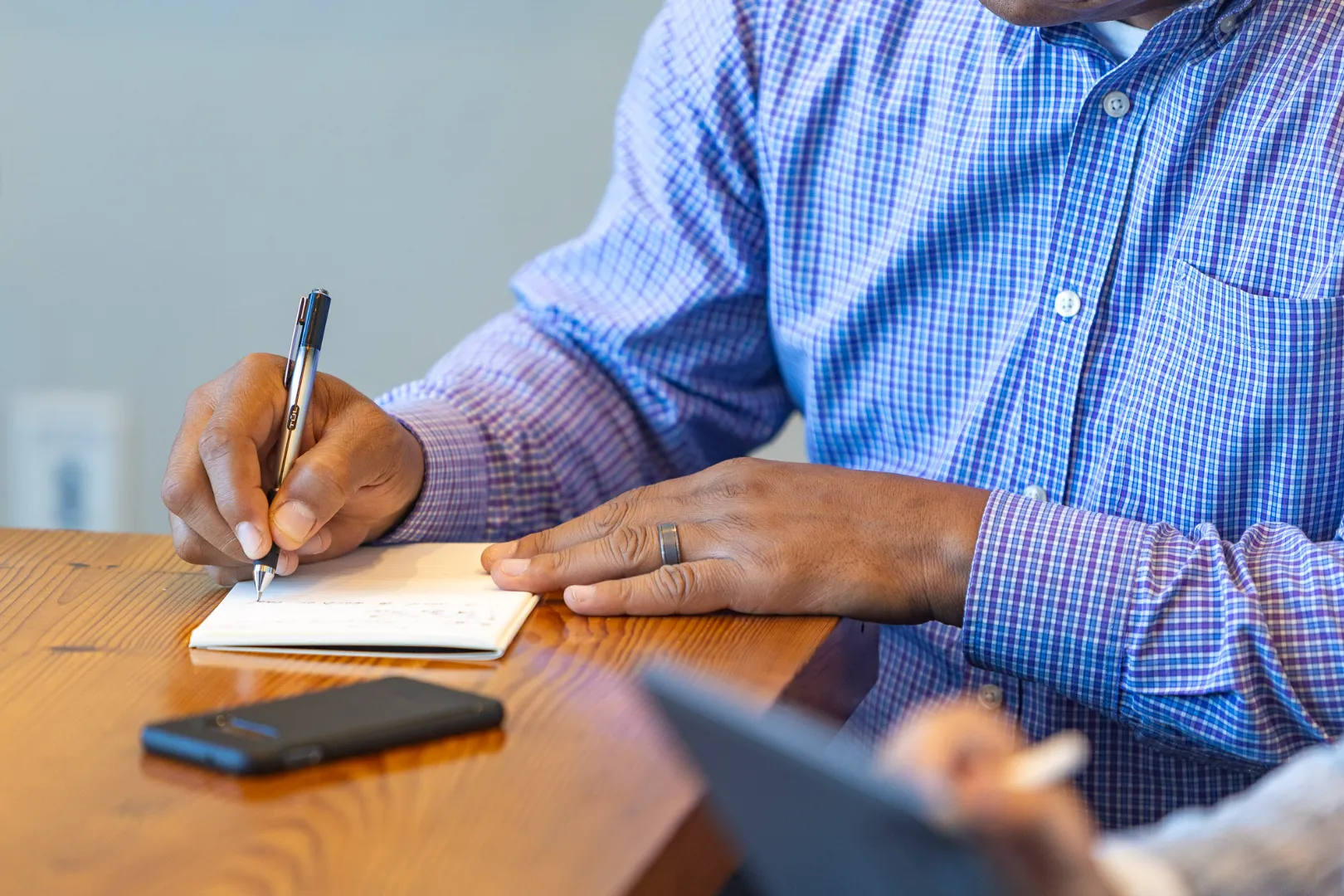 Person in a blue checkered shirt writing on a notepad with a pen at a wooden table, with a smartphone nearby.