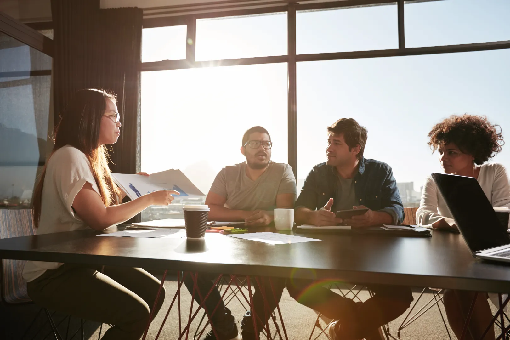 Four colleagues having a discussion around a table in a sunlit office with documents and coffee cups.