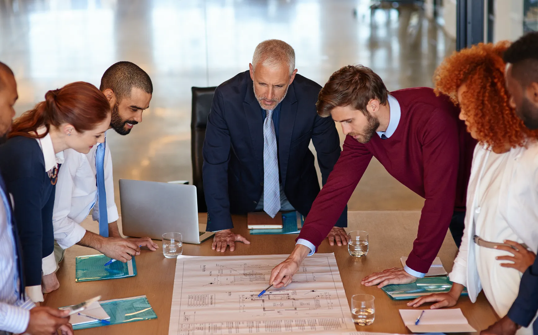 A diverse group of six businesspeople gathered around a table reviewing architectural blueprints during a meeting.