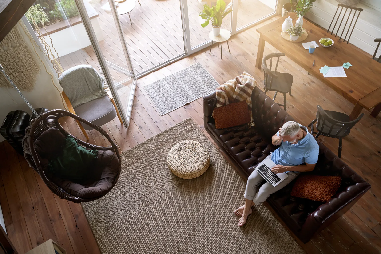 Man with gray hair sitting barefoot on a brown leather sofa using a laptop in a cozy living room with natural light and wooden floors.