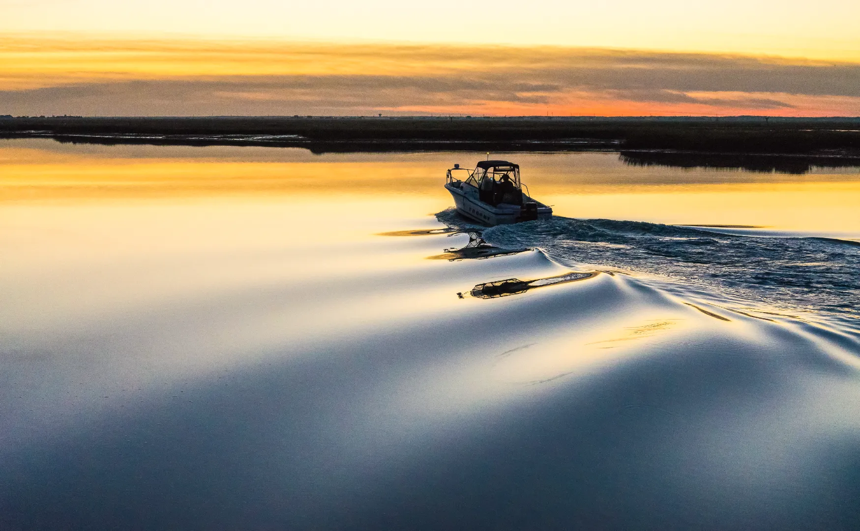 Small motorboat creating ripples on calm water during a golden sunset.