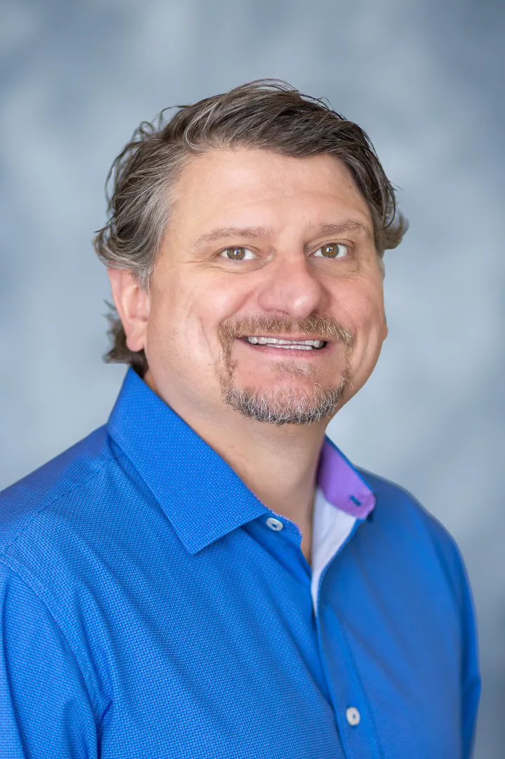 Smiling man with short wavy gray hair and a goatee wearing a blue button-up shirt against a blurred gray background.