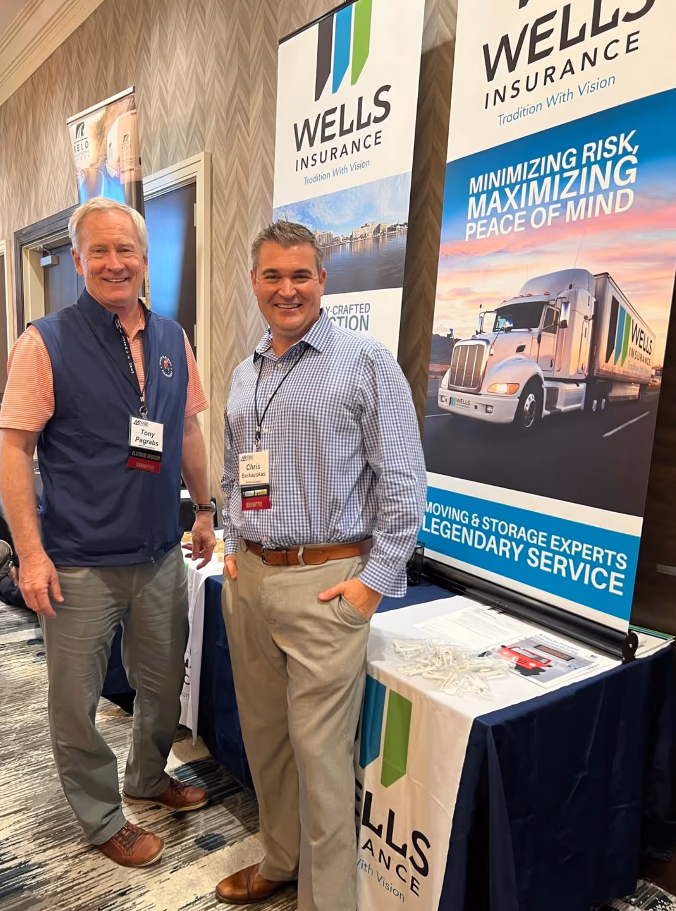 Two smiling men standing in front of Wells Insurance banners at a conference booth.