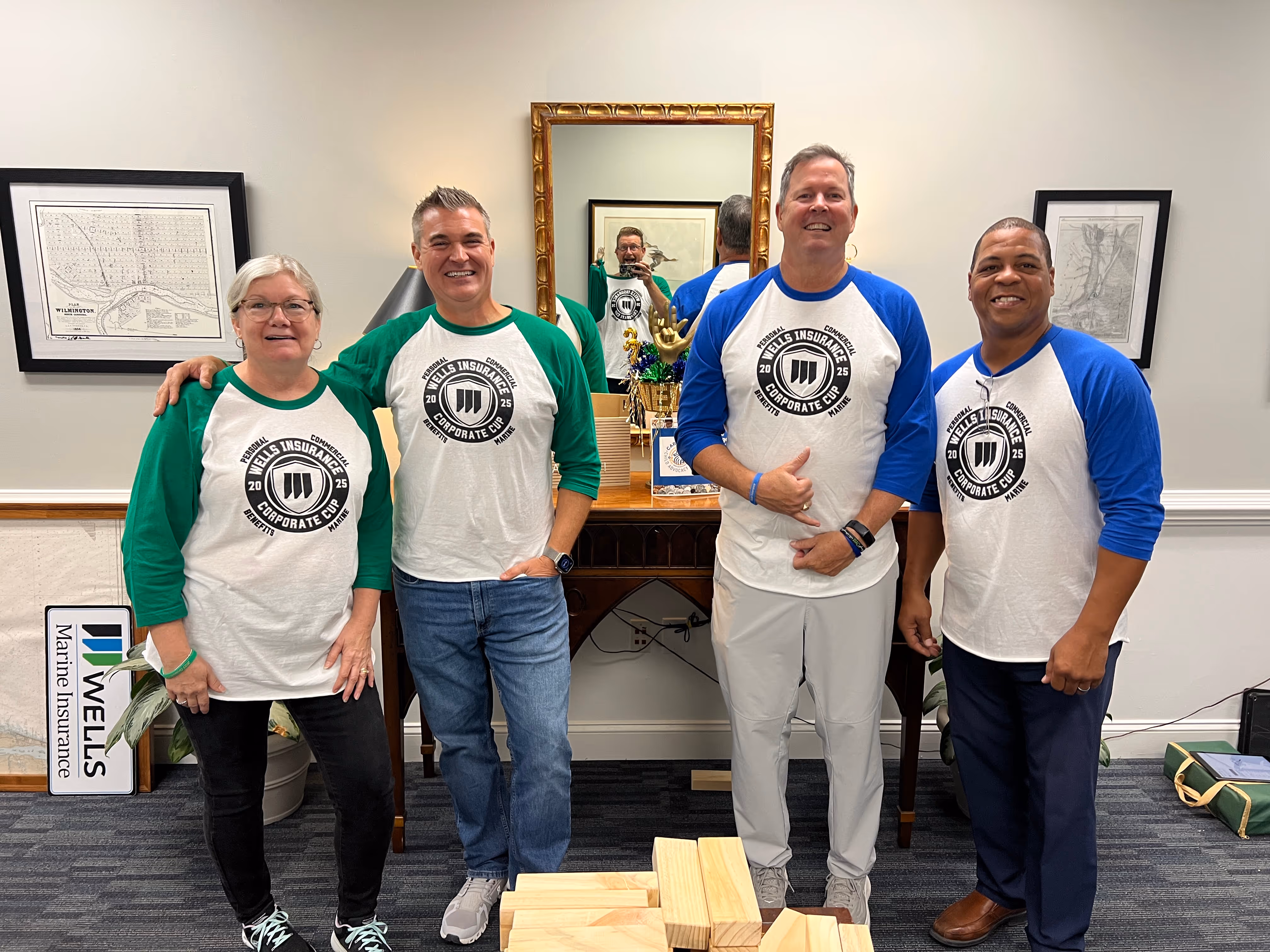 Four people smiling indoors wearing matching Wells Insurance Corporate Cup baseball-style shirts with green and blue sleeves.