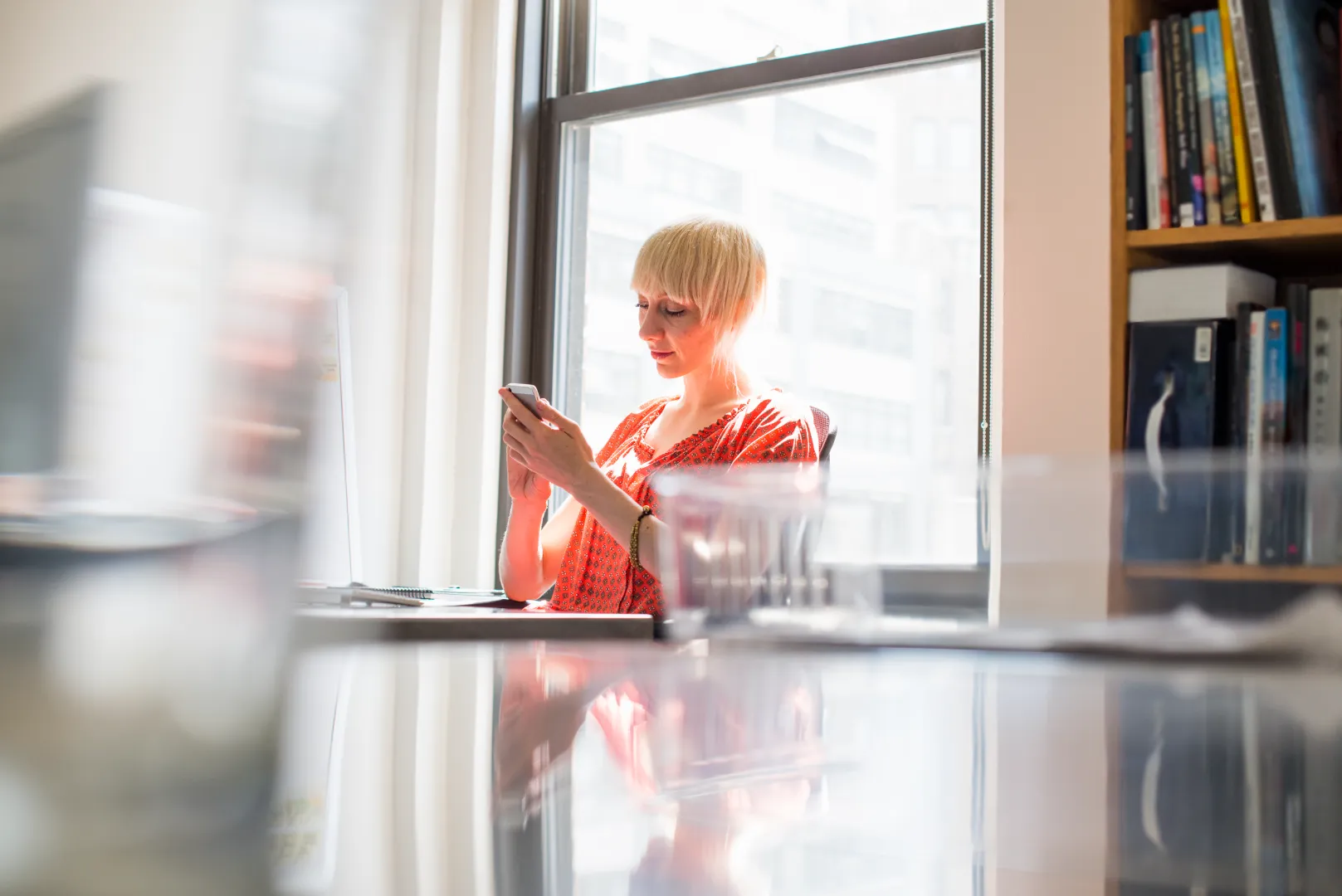 Young woman in orange shirt sitting at a desk by a window, focused on her smartphone.