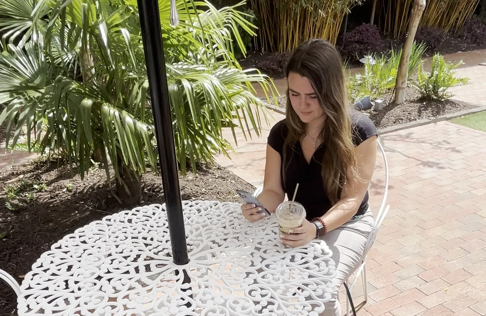 Woman sitting outdoors at a white ornate metal table holding a smartphone and iced coffee, surrounded by greenery.