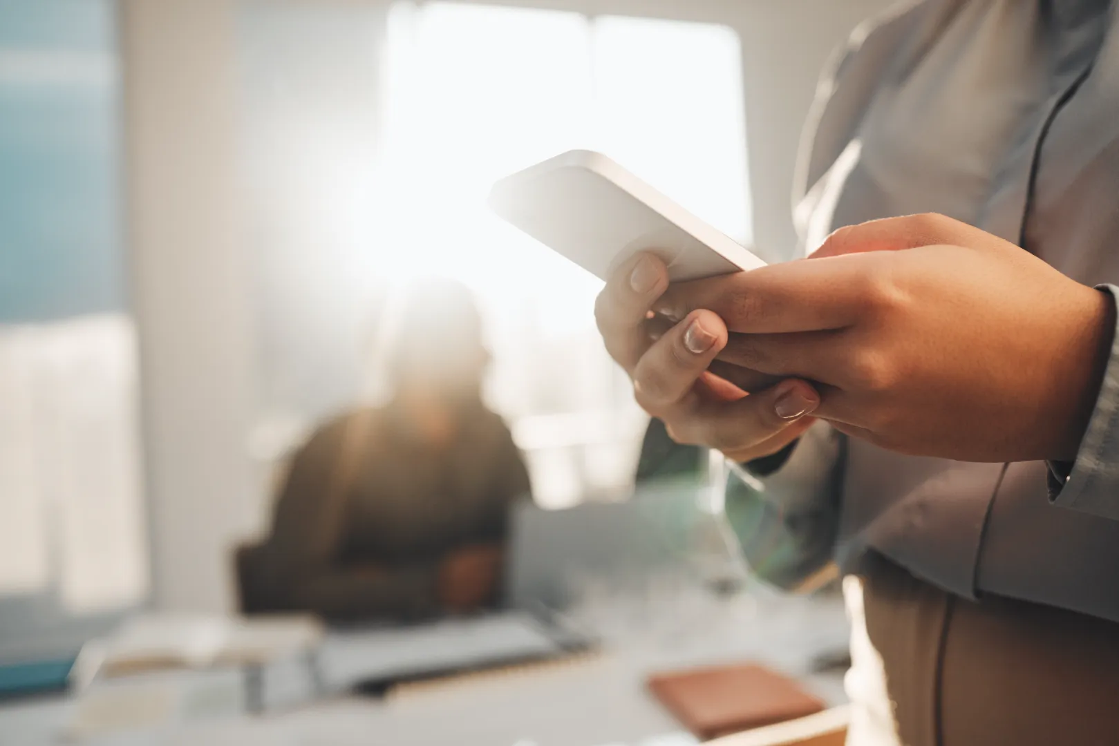 Person holding and using a smartphone in a sunlit office with another person blurred in the background.