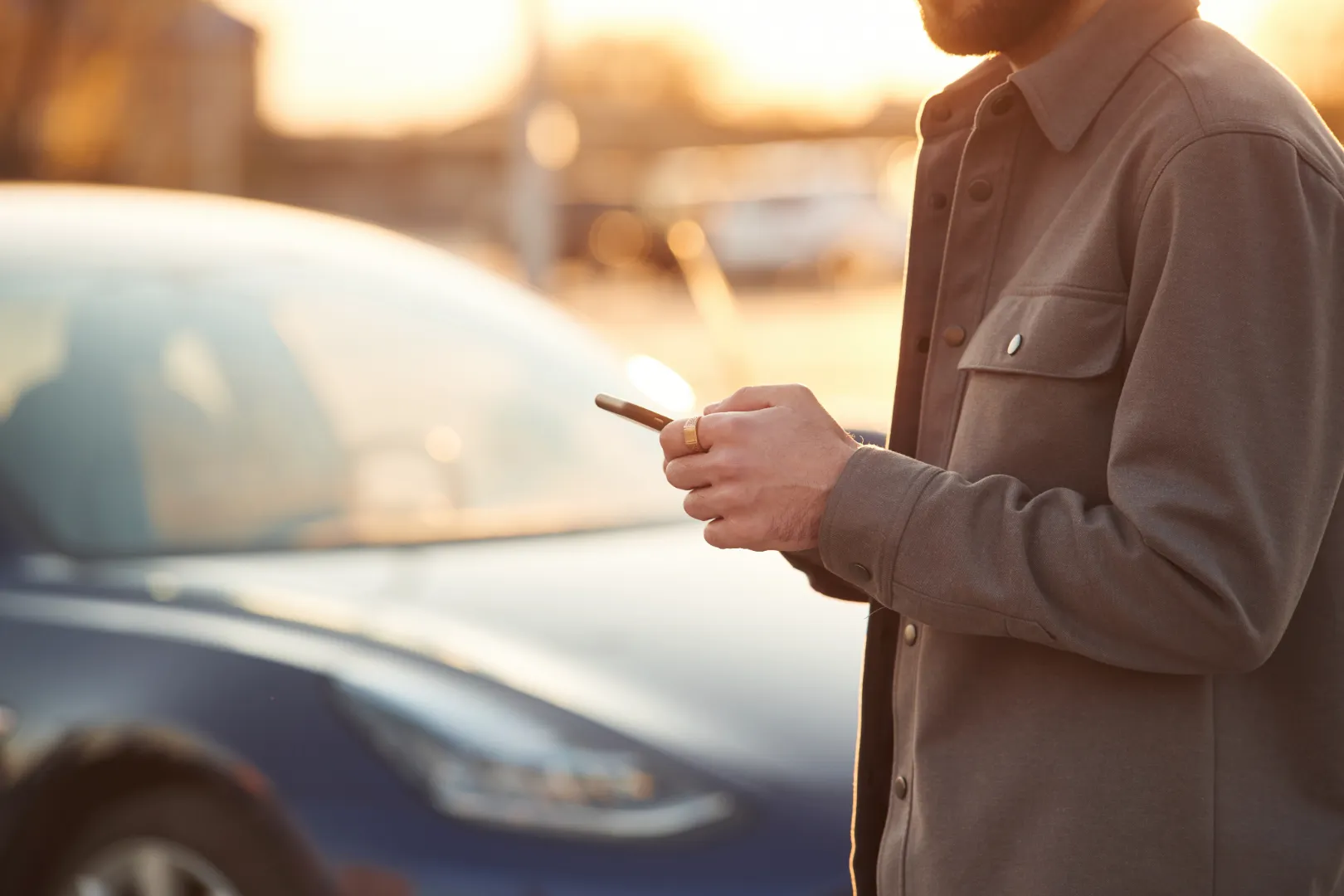 Man in brown jacket using smartphone with a parked blue car in the background during sunset.
