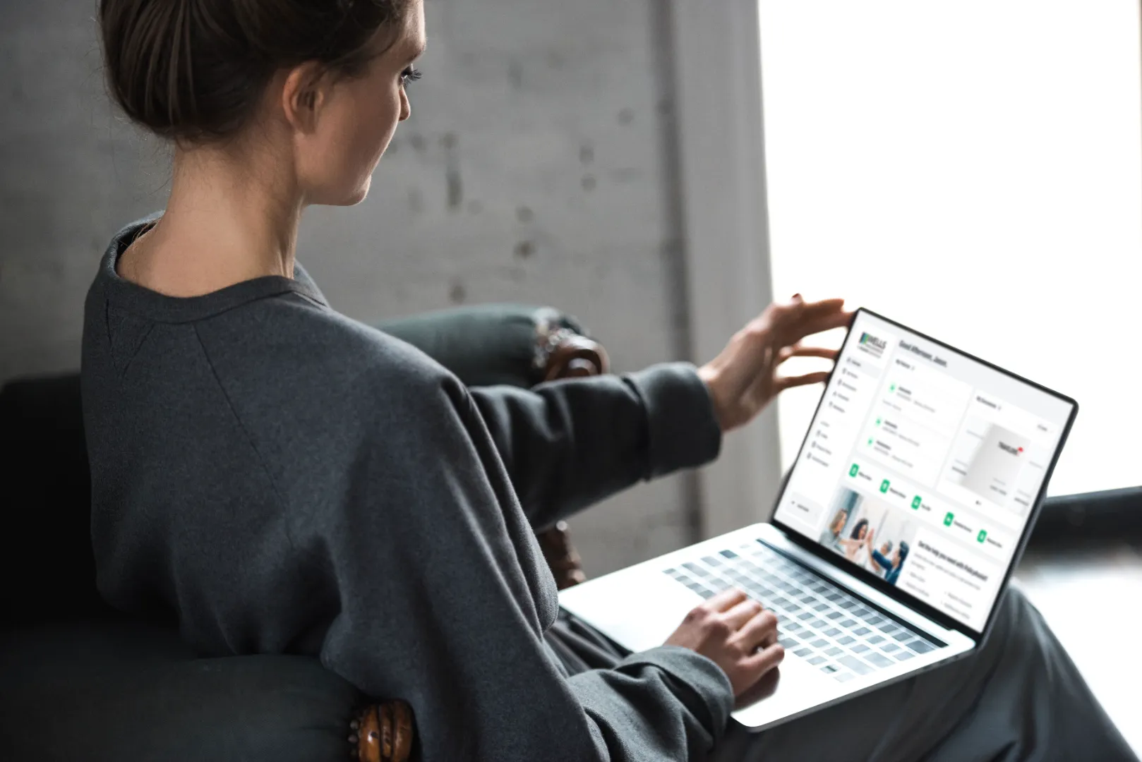 Person wearing a dark sweater seated in an armchair, using a laptop displaying a dashboard interface.