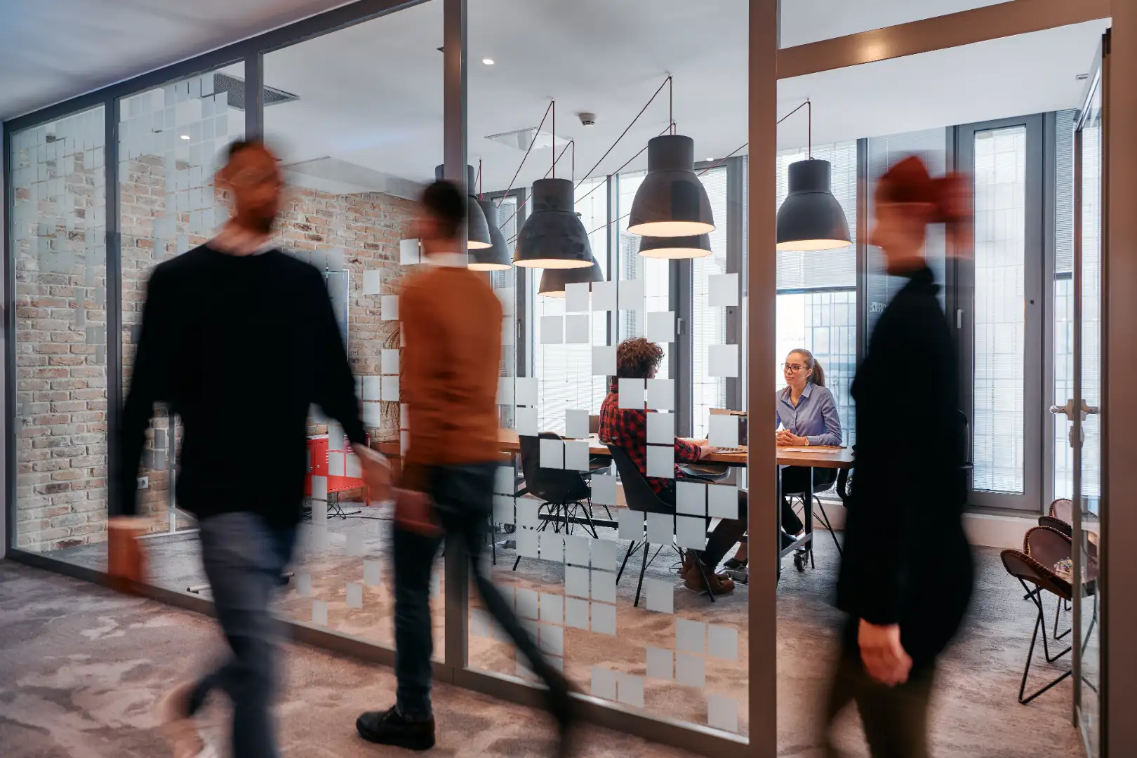 Office scene with blurred figures walking past a glass-walled meeting room where two people are seated and talking.