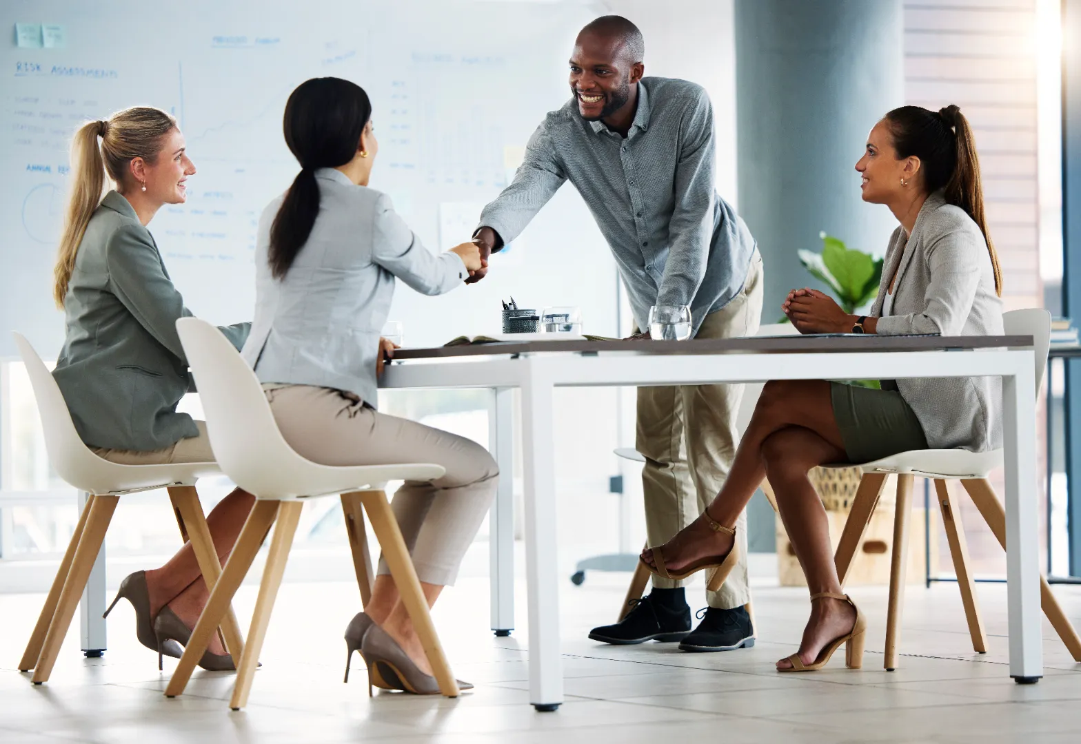 Three women seated around a table with a man standing and shaking hands with one of them in a modern office.