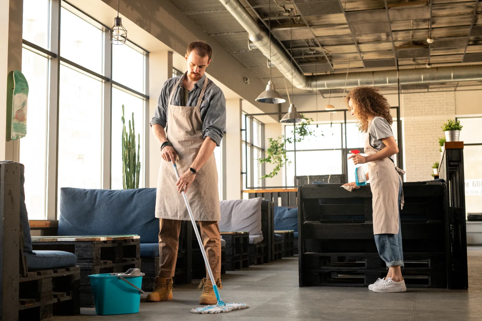 Two people wearing aprons cleaning a modern cafe interior, one mopping the floor and the other wiping a bench with spray.