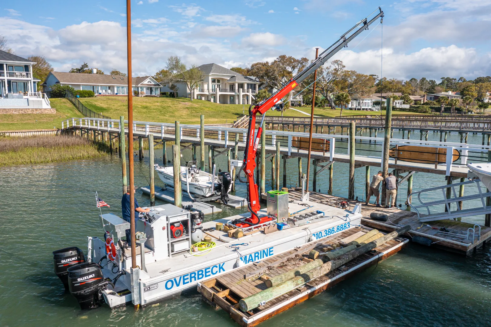 Commercial marine workboat with a red crane lifting wooden poles near a dock by waterfront houses.