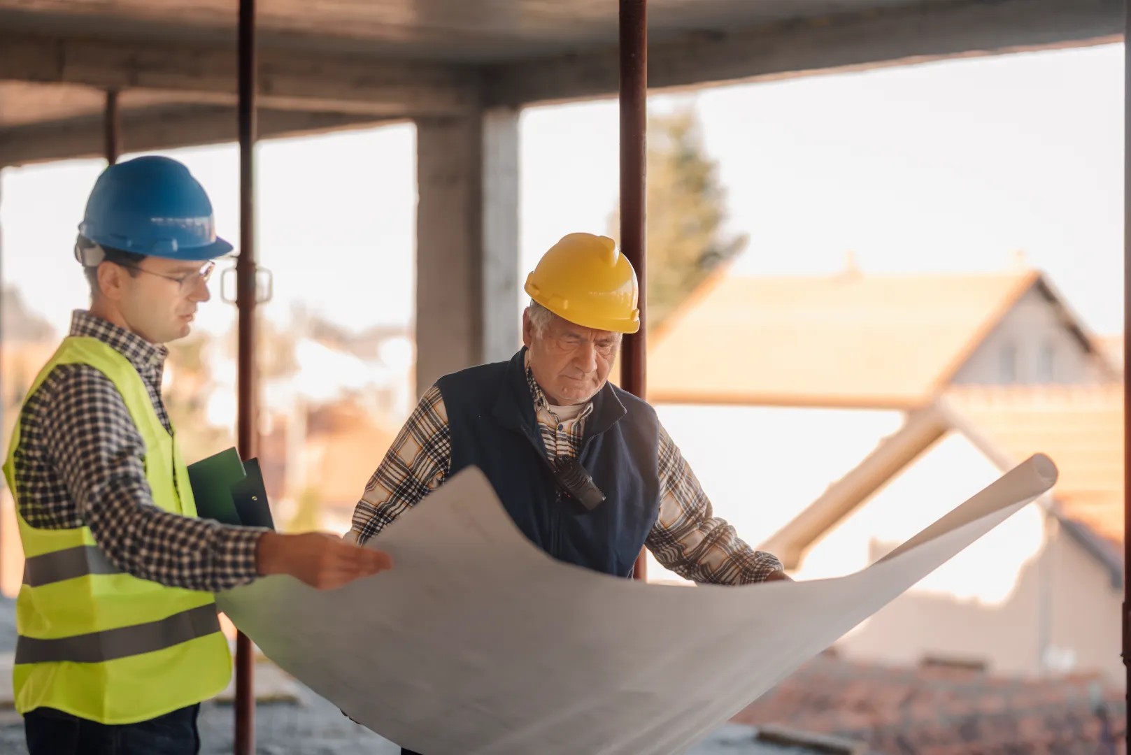 Two construction workers wearing helmets and safety gear reviewing a large blueprint on a building site.