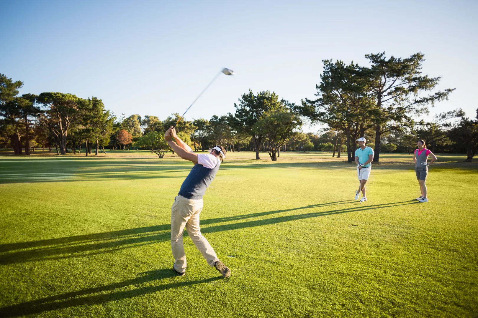 Man swinging a golf club on a sunny green golf course while two others watch nearby.
