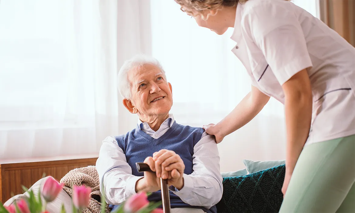 Smiling elderly man sitting with a cane looking up at a caregiver who is gently touching his shoulder.