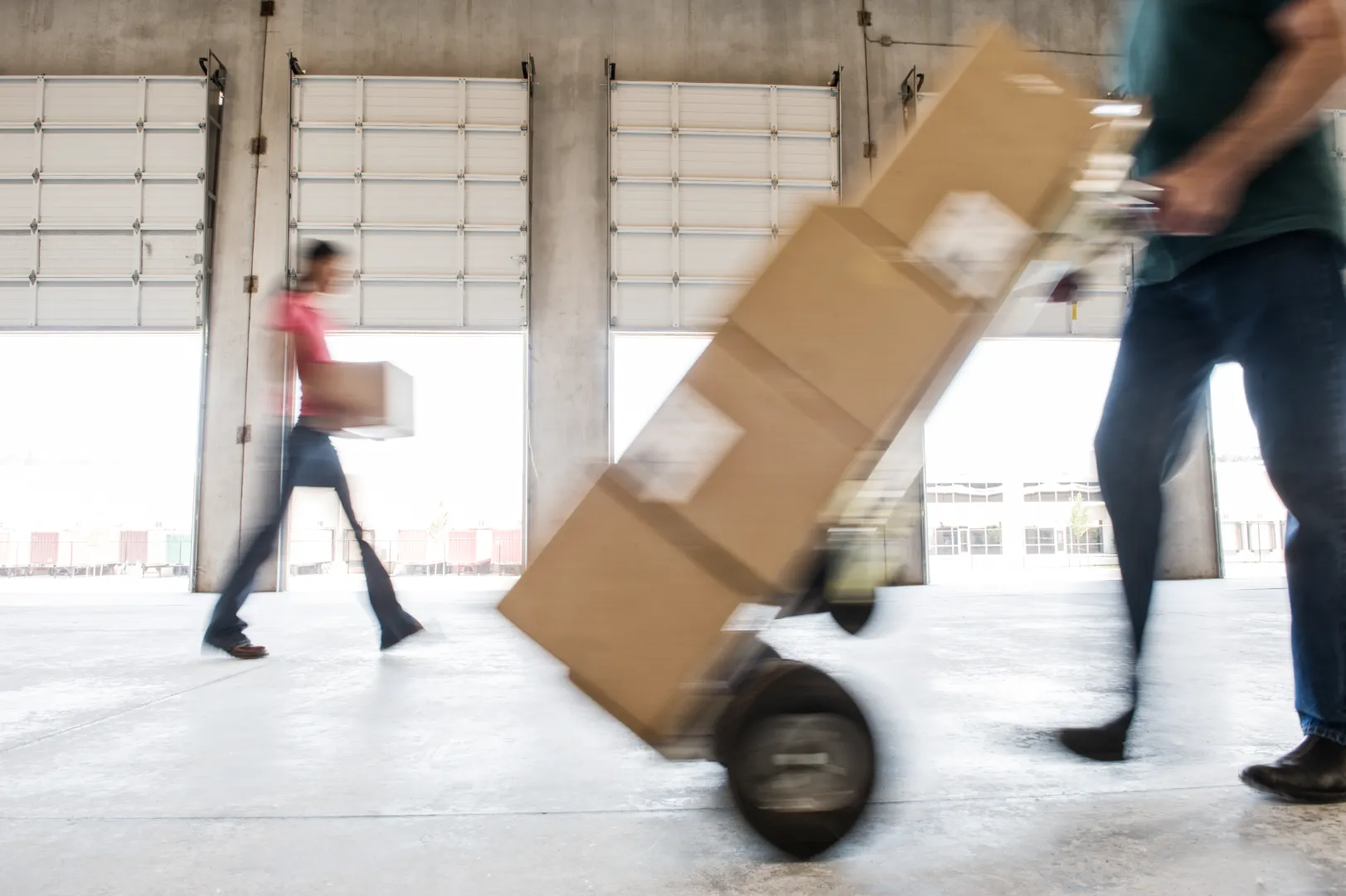 Blurry image of two movers in a warehouse, one pushing a dolly with stacked boxes and another carrying a box.