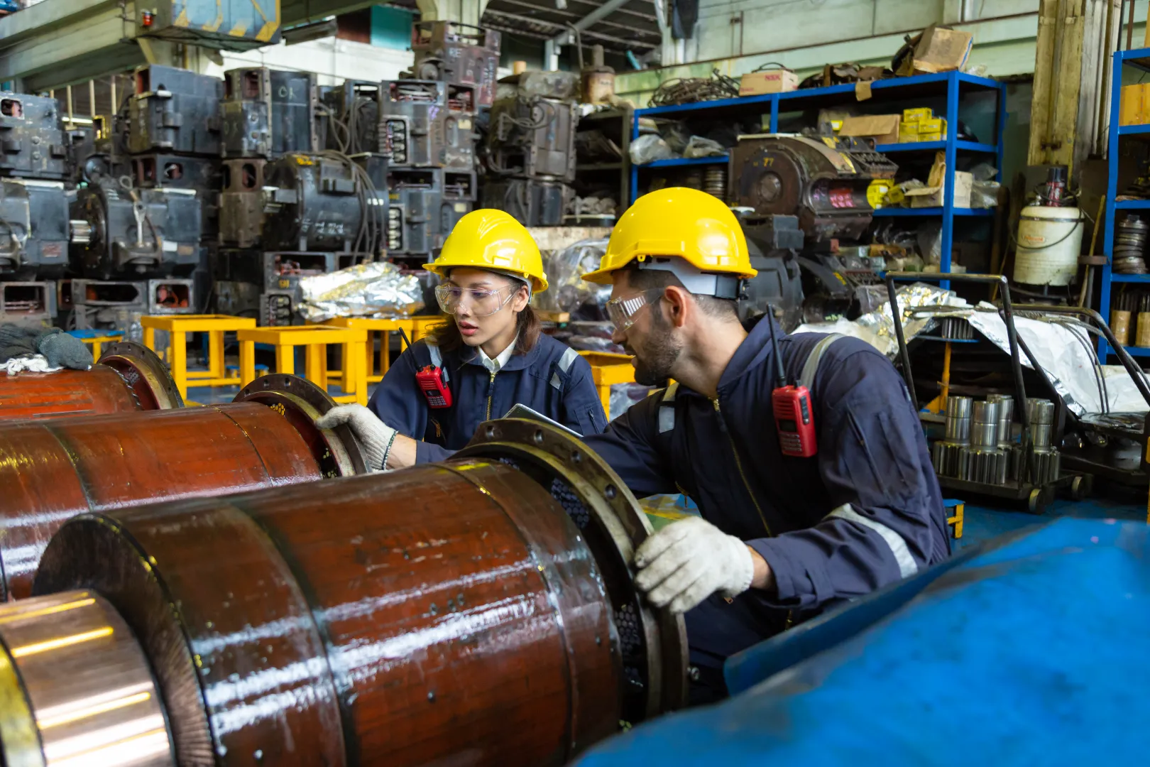 Two industrial workers wearing yellow hard hats and protective glasses inspecting large machinery parts in a factory.