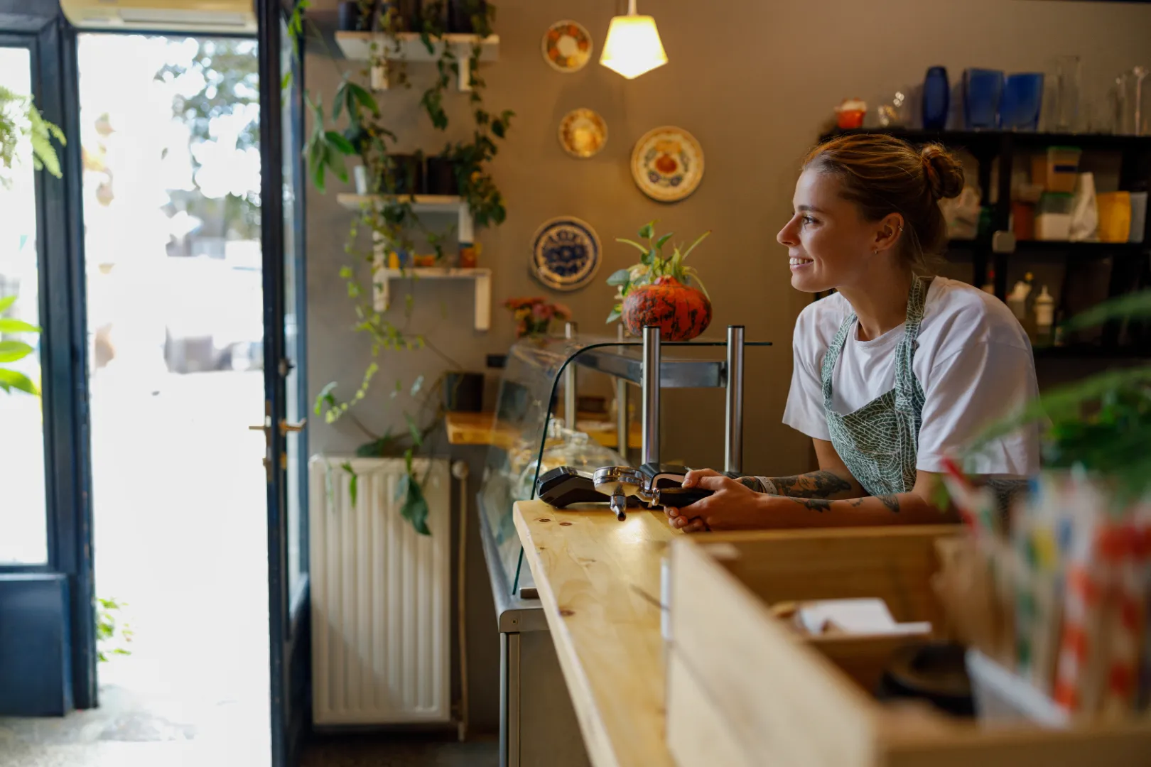 Smiling woman in apron leaning on a wooden counter inside a cozy café with plants and decorative plates on the wall.