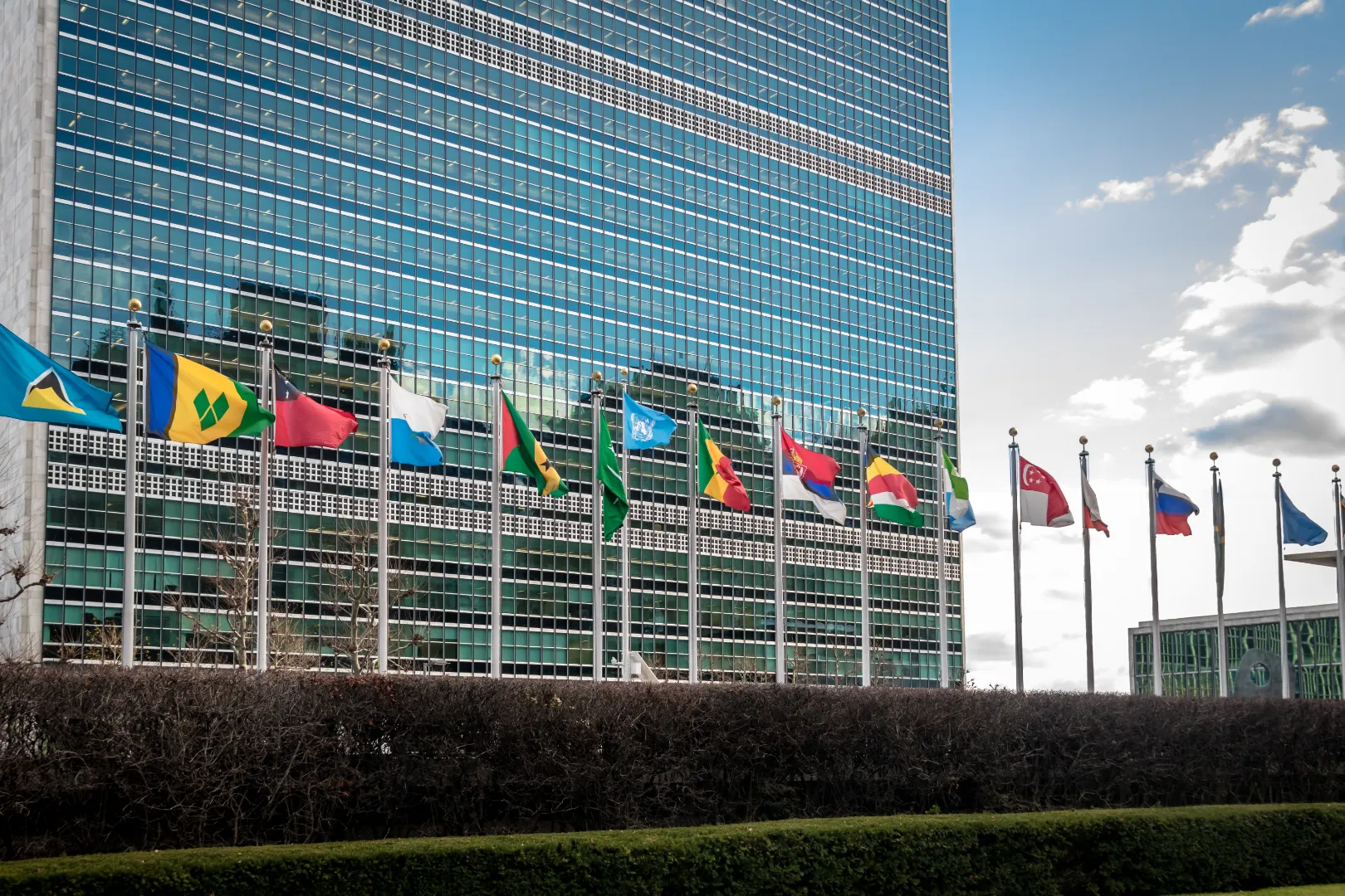 Multiple international flags flying on flagpoles in front of a glass-paneled building under a partly cloudy sky.