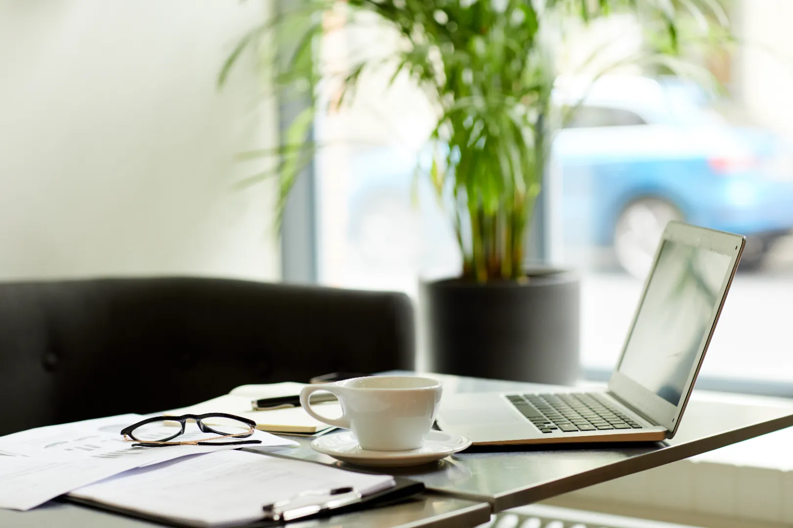 Workspace with an open laptop, white coffee cup on saucer, eyeglasses, papers, and a pen on a table near a black chair and a potted plant.
