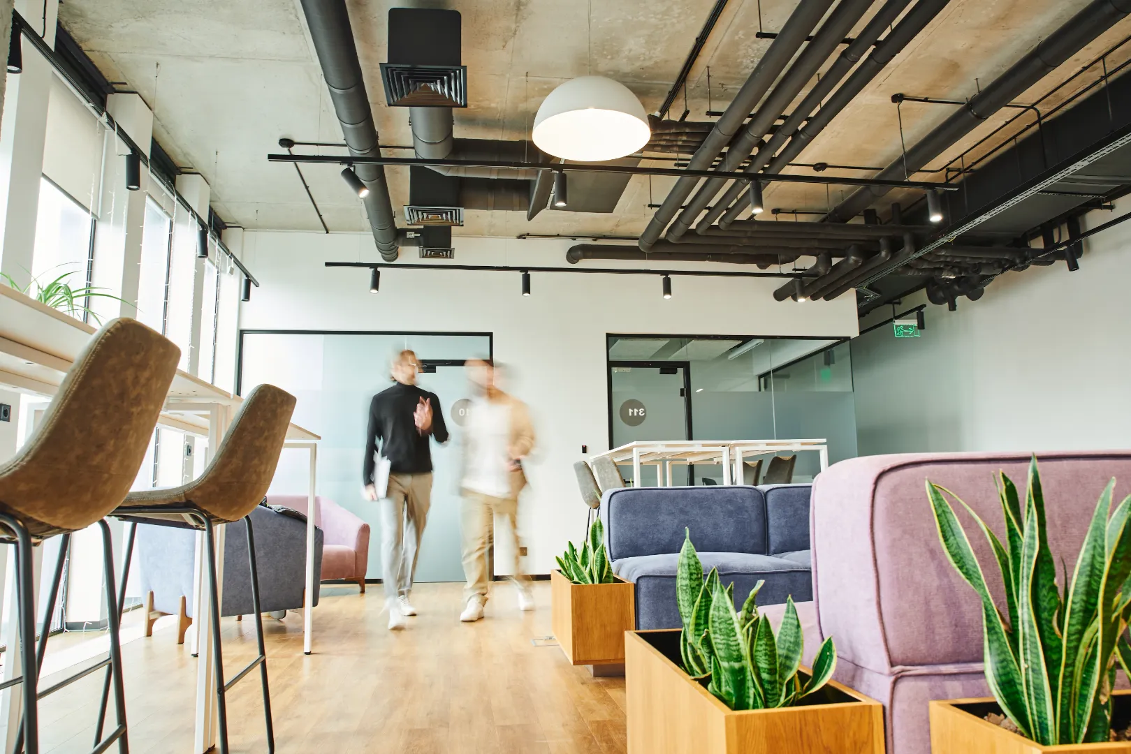 Modern office lounge with purple and blue sofas, wooden planters with green plants, high chairs, and two blurred figures walking.