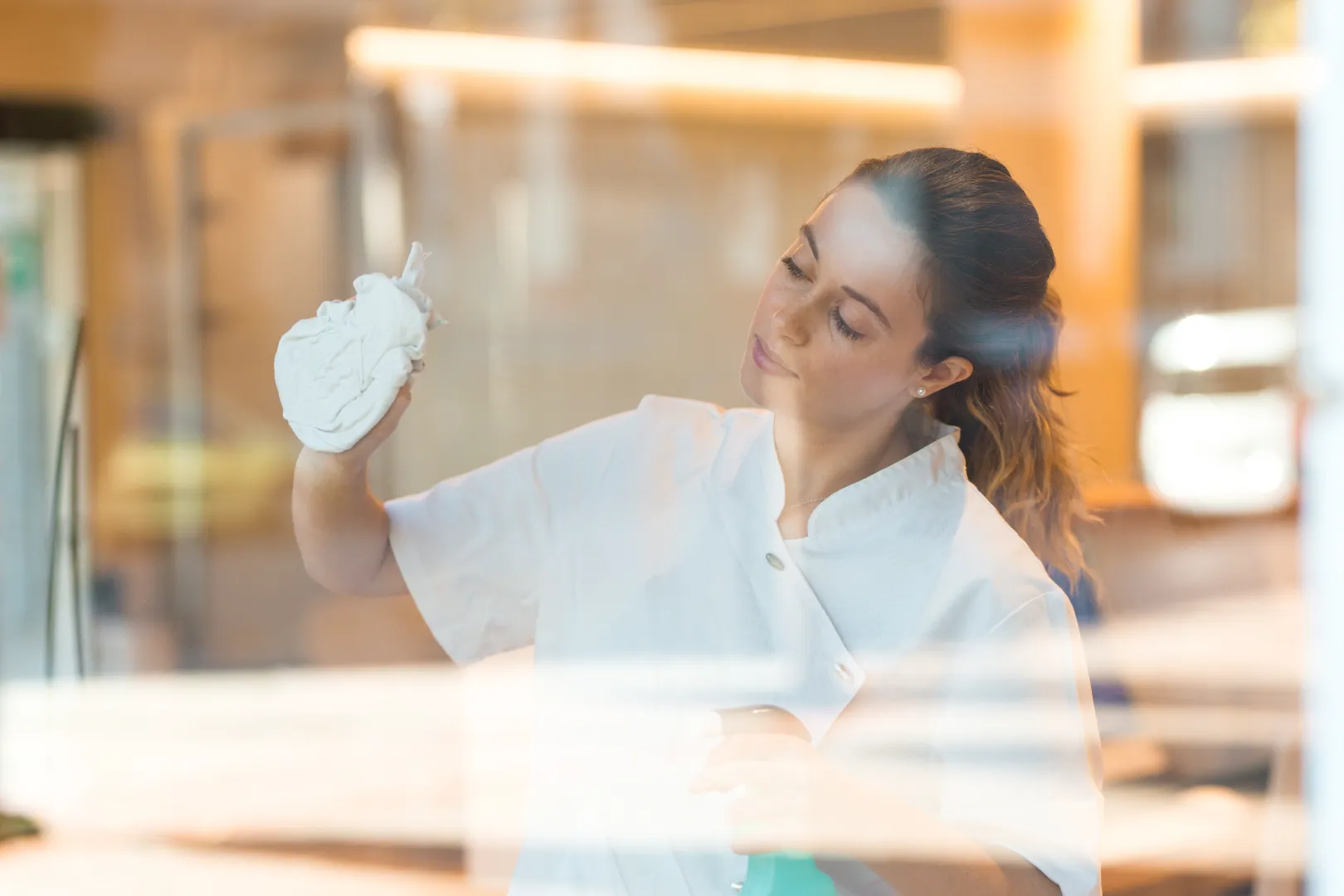 Woman in white shirt cleaning a glass surface with a cloth and spray bottle indoors.