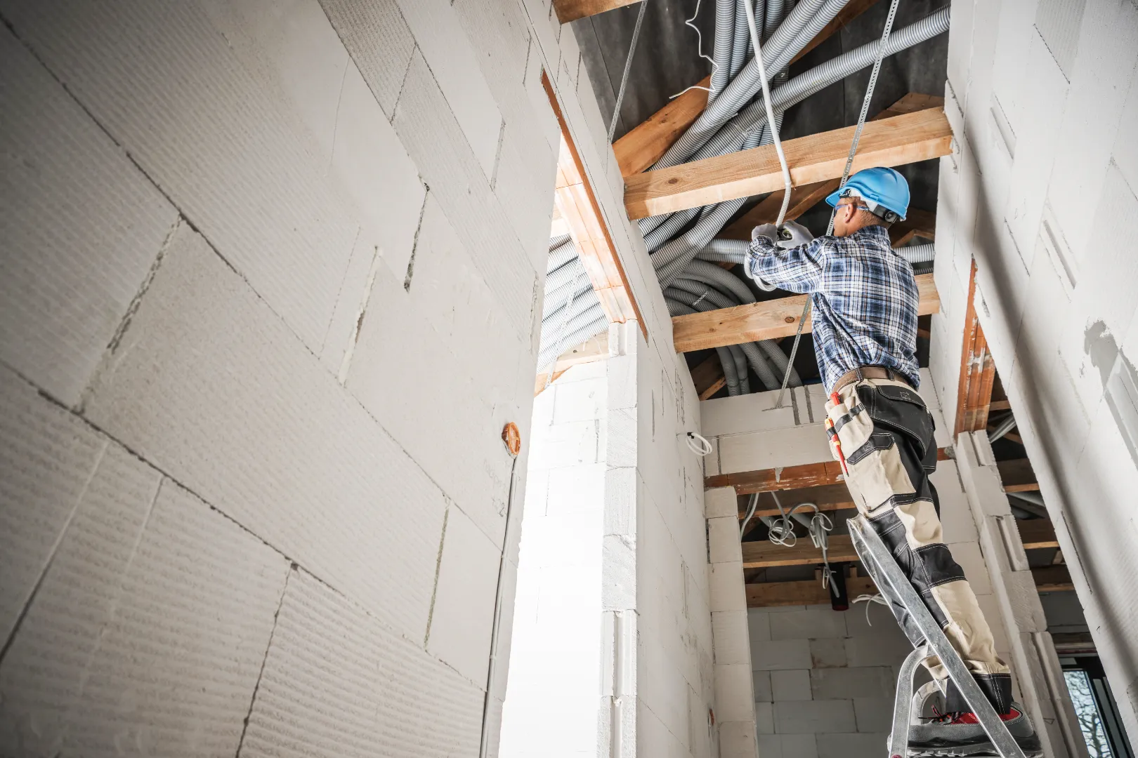 Construction worker in a blue helmet standing on a ladder installing electrical cables in an unfinished building.