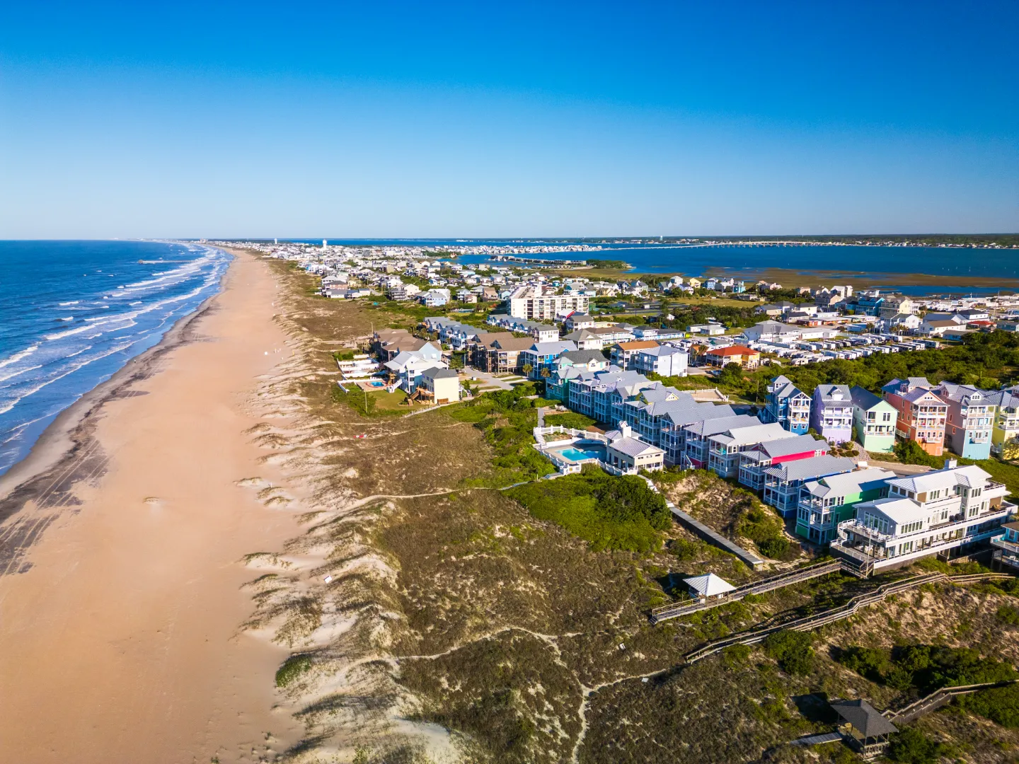 Aerial view of a coastal beachfront community with houses, sandy beach, ocean waves, and a clear blue sky.