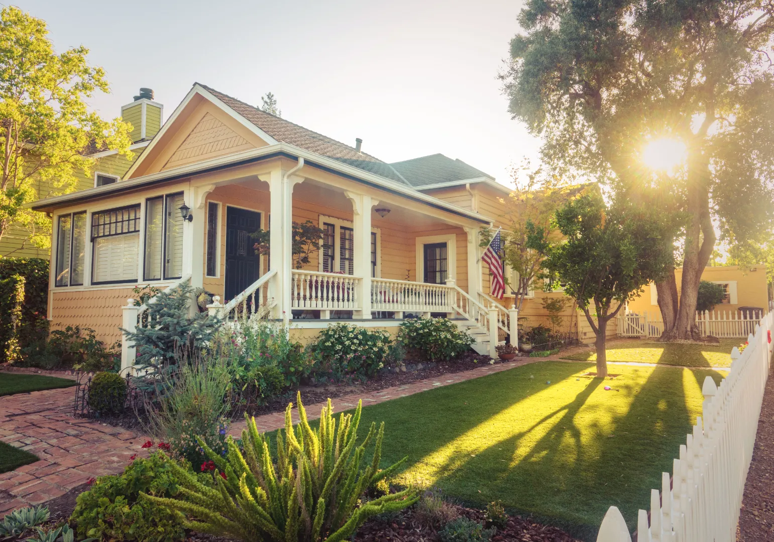 Yellow house with a white porch railing, American flag, garden flowers, green lawn, and sunlight filtering through trees.