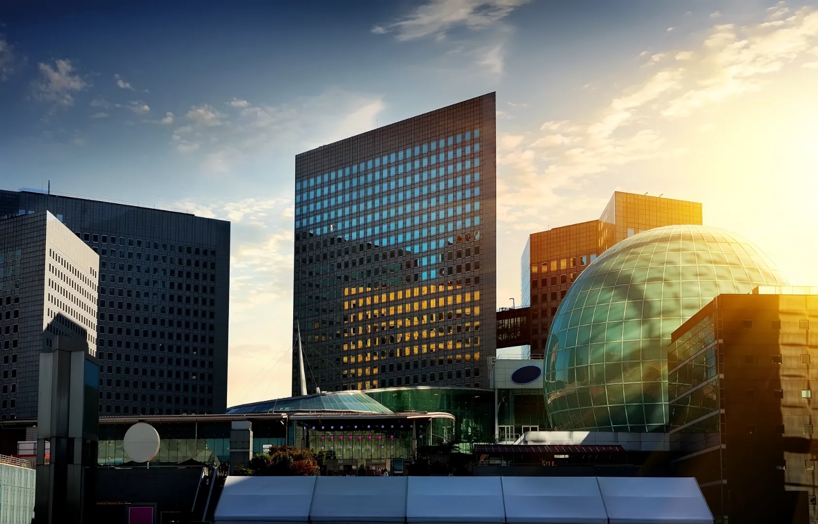 Modern cityscape with glass skyscrapers and a large glass dome reflecting sunlight at sunset.