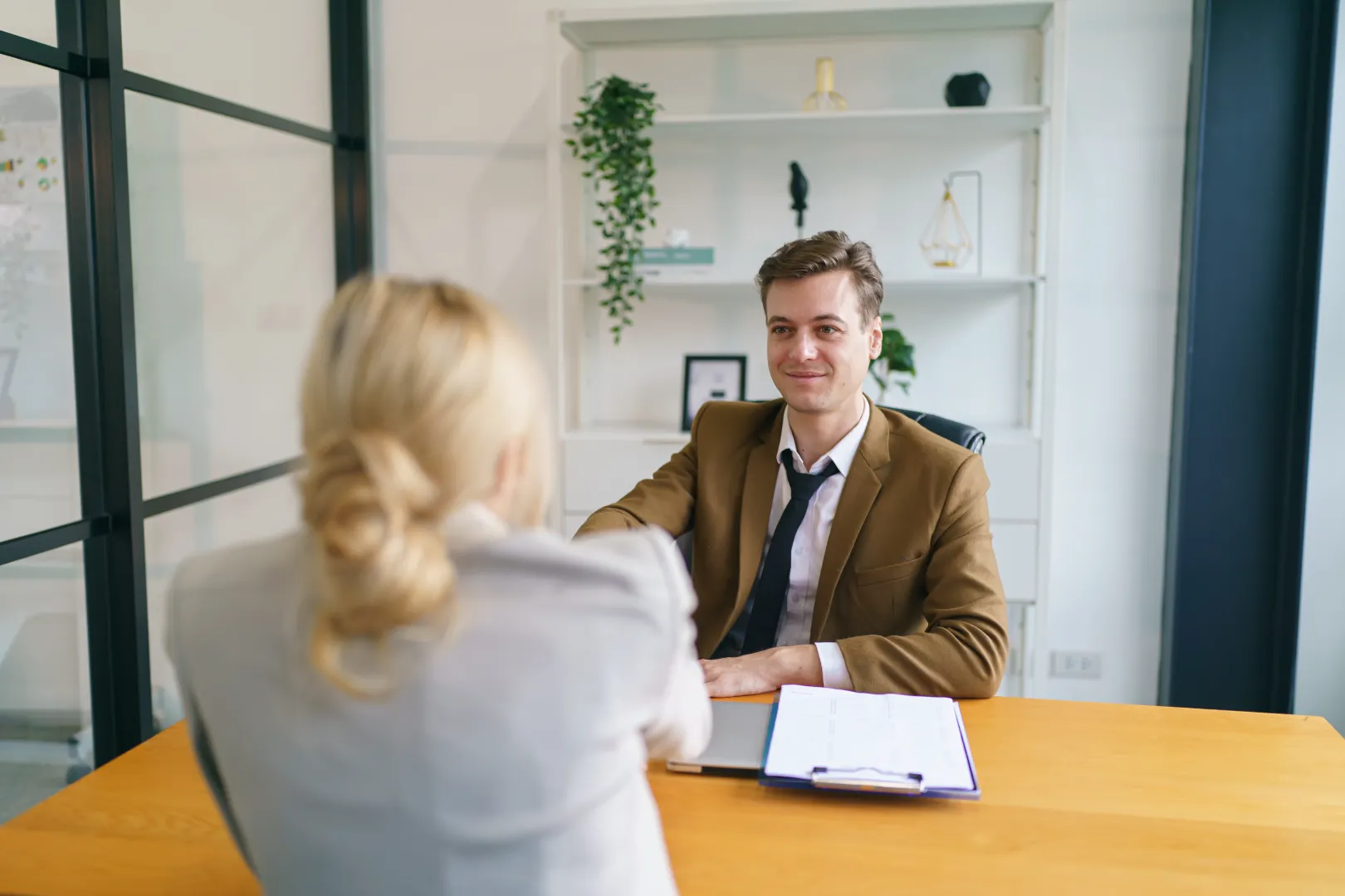 Young man in a brown blazer shaking hands with a woman in a grey jacket during a meeting at an office desk.