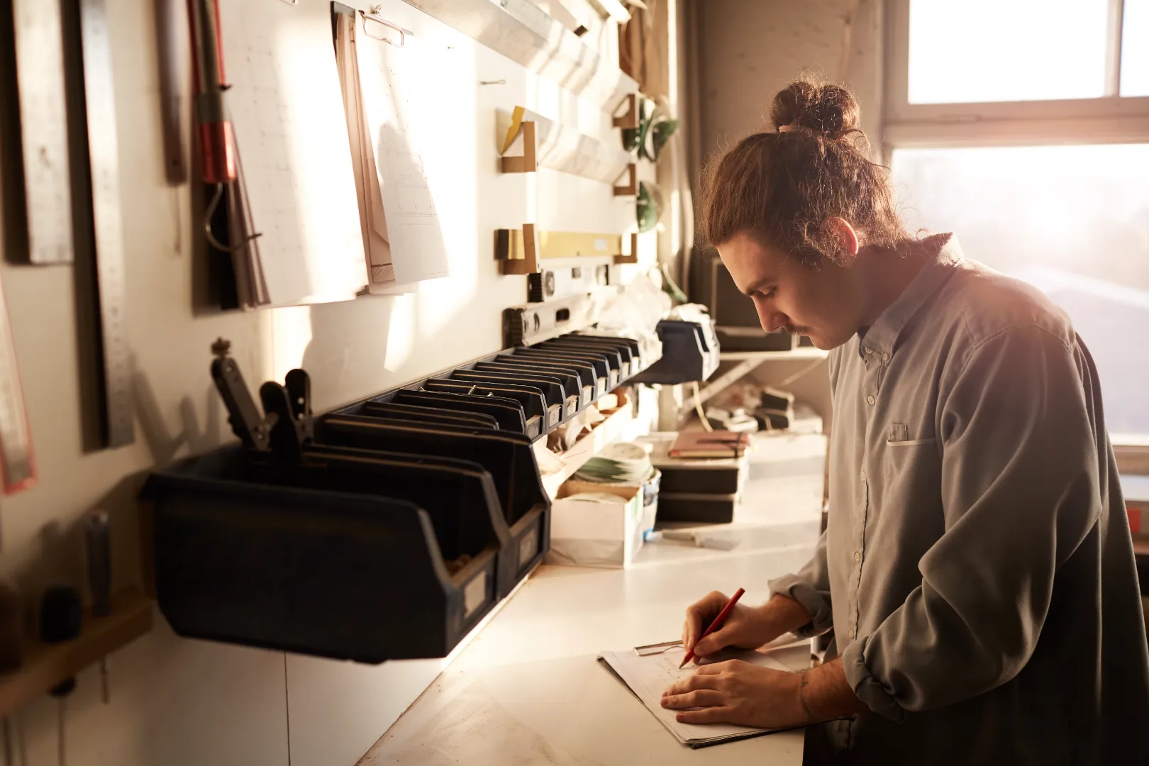 Man with a bun hairstyle writing or drawing on a clipboard at a well-organized workspace bathed in warm sunlight.
