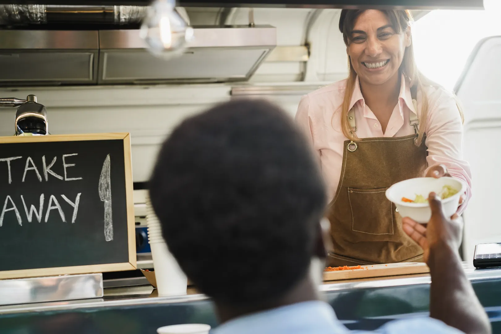 A smiling woman in a brown apron hands a takeout bowl of food to a customer at a food service counter.