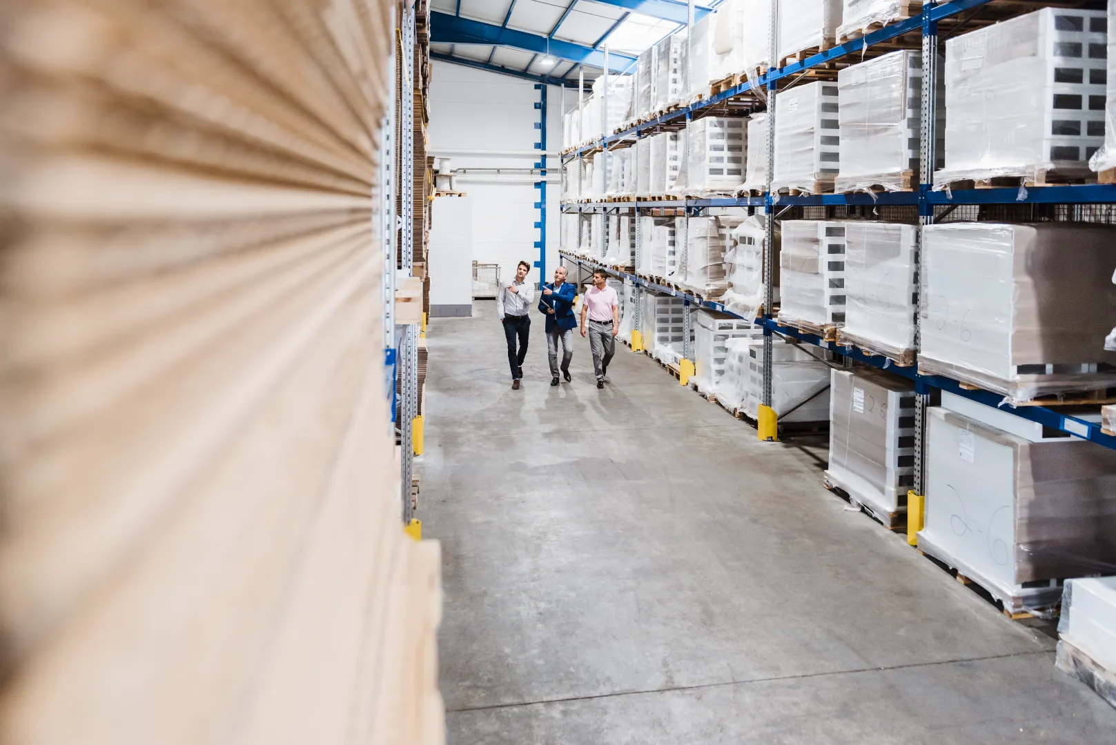 Three men walking and discussing inside a warehouse aisle with stacked, wrapped pallets on metal shelves.