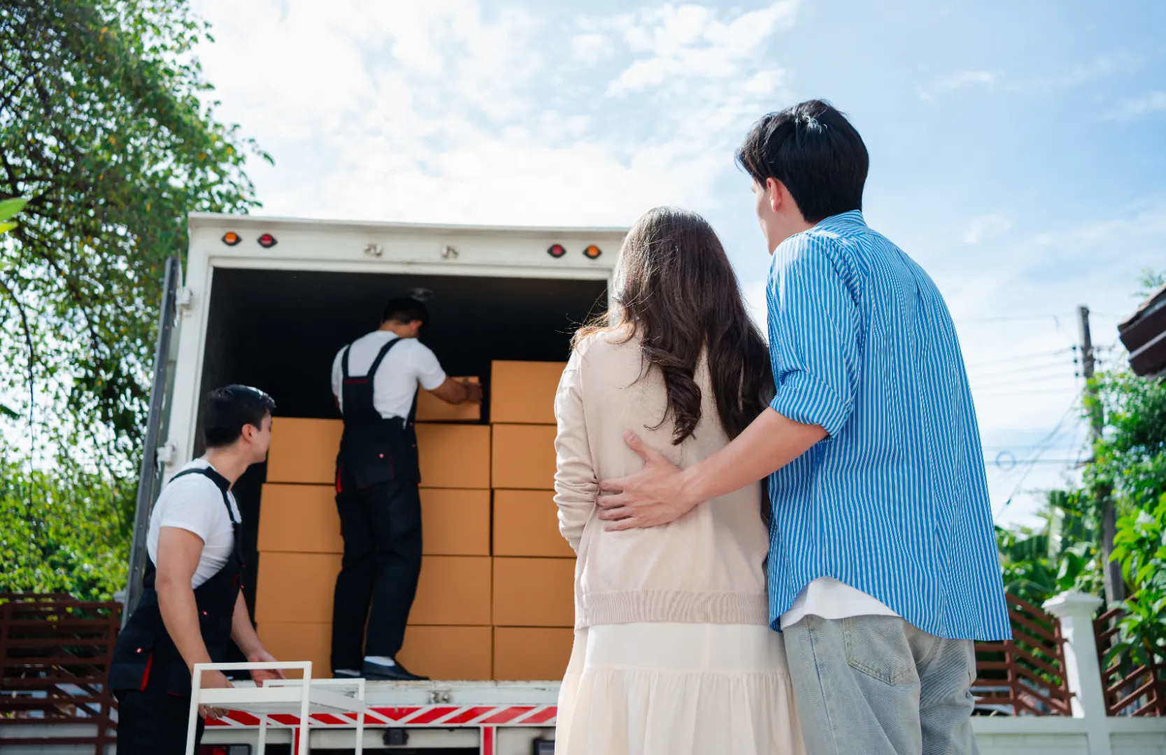A couple standing outside watching two movers loading stacked cardboard boxes into a truck on a sunny day.